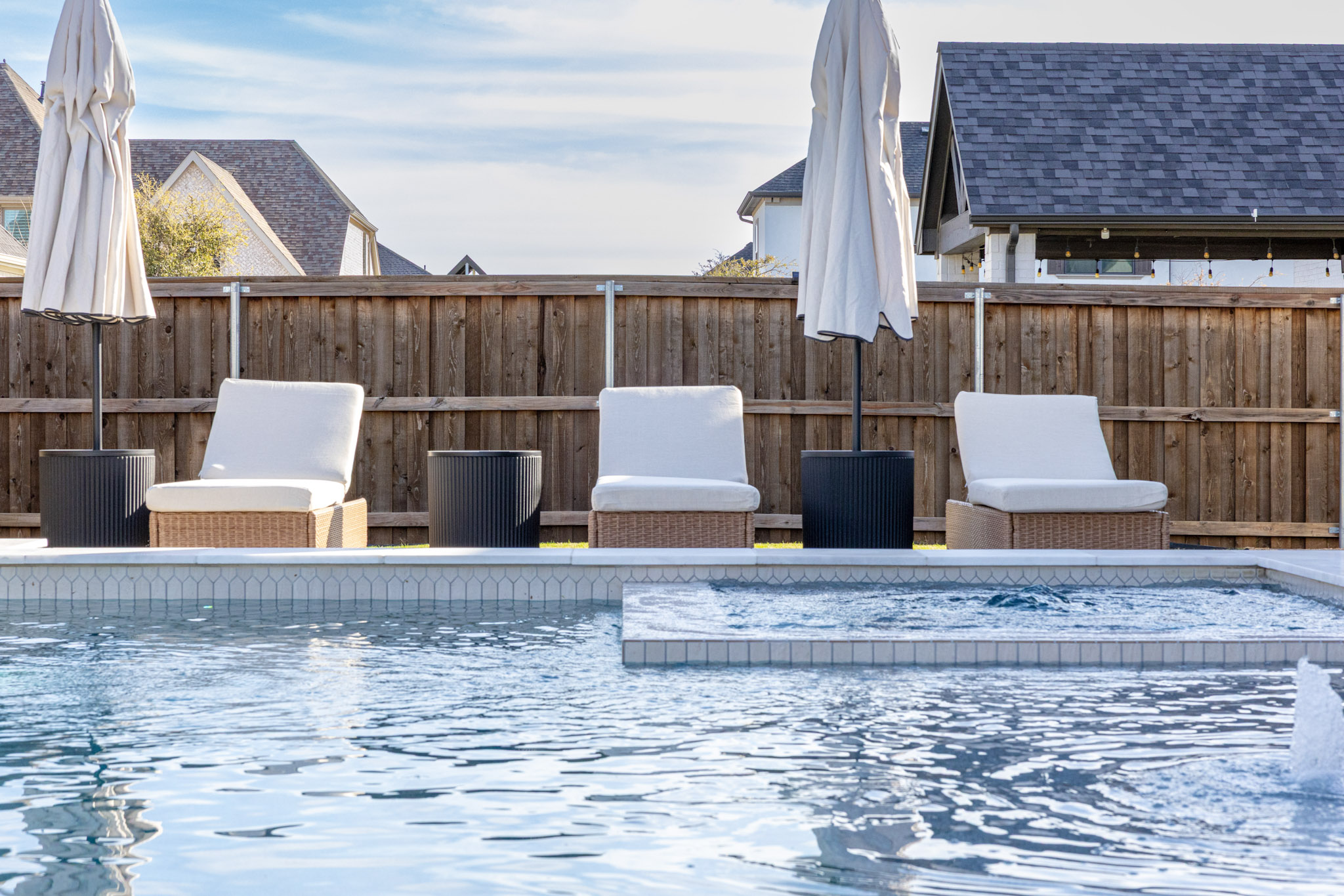 Square pool spa with three cushioned lounge chairs and closed umbrellas behind it.