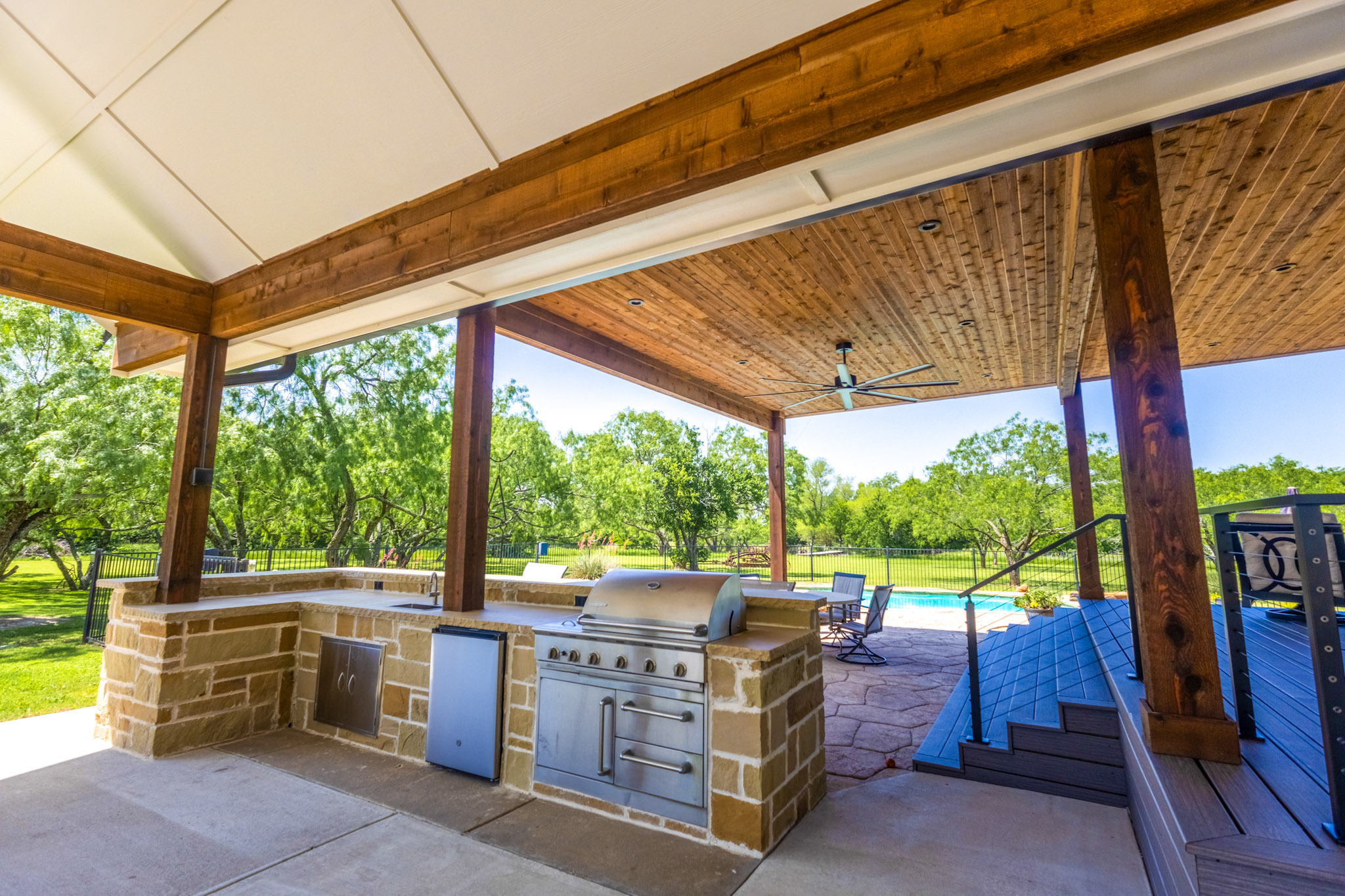Vaulted covered patio with exposed wood beams, ceiling fan and stone outdoor kitchen.