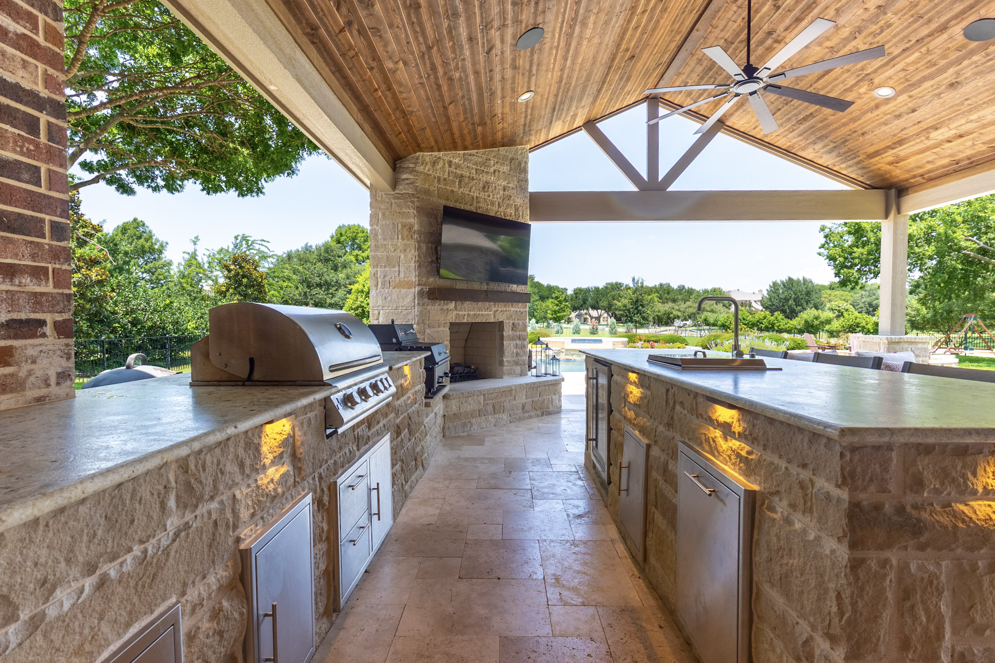 Stone outdoor kitchen with built-in grills, bar island, fireplace, and vaulted wood patio cover.