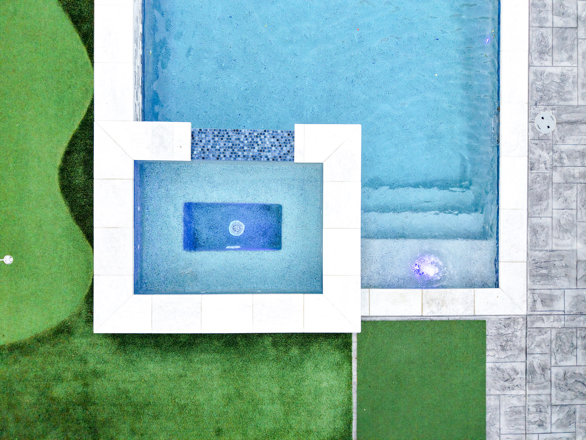 Aerial detail of raised square spa beside pool steps with white coping and artificial turf.