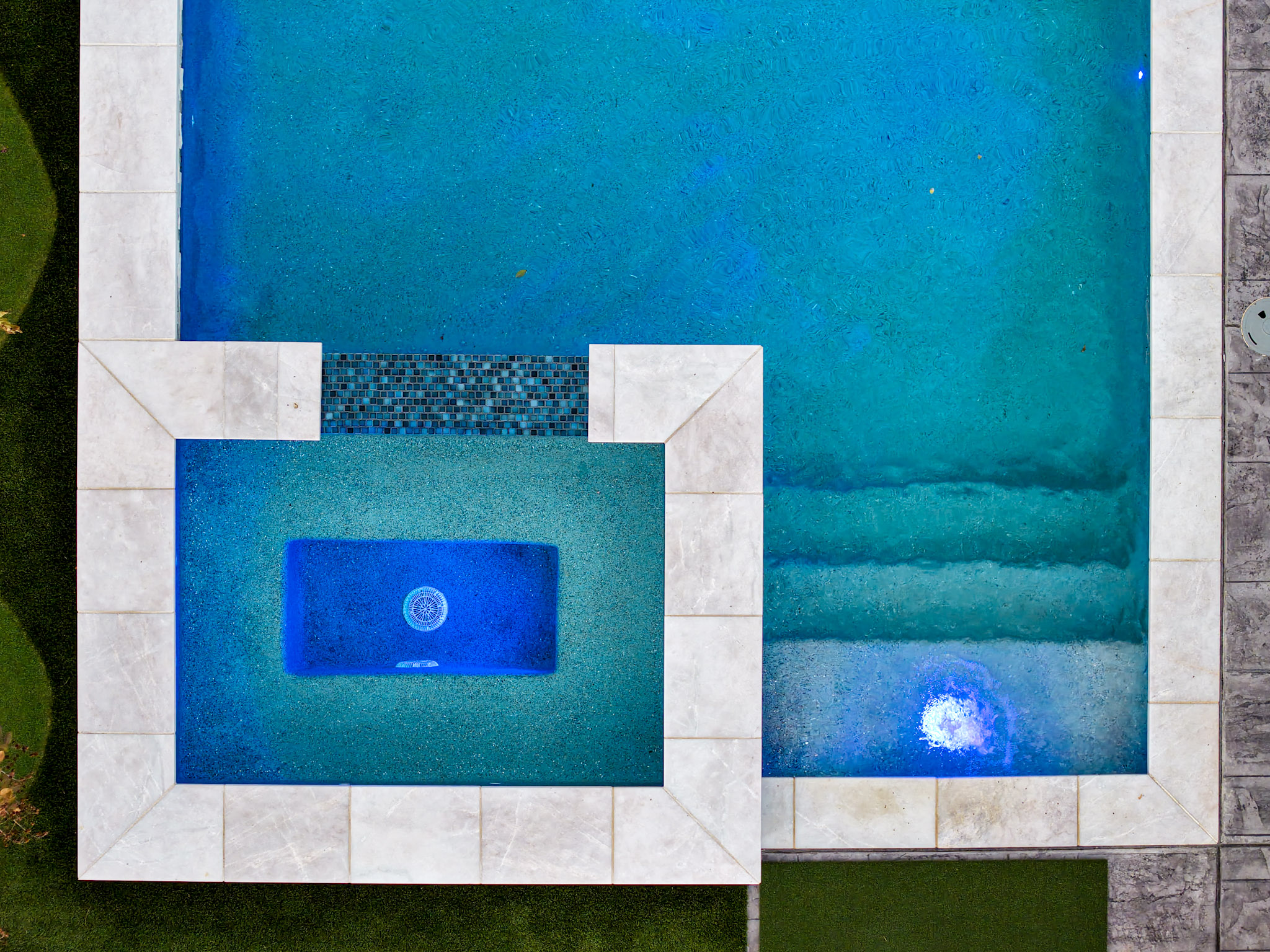 Top-down view of square raised spa beside a rectangular pool with entry steps.
