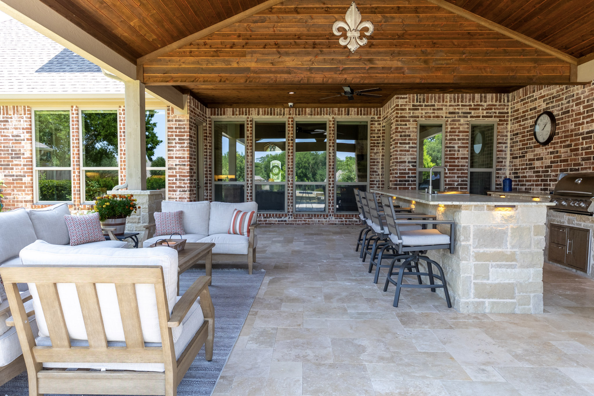 Covered patio with lounge furniture, stone outdoor kitchen, bar seating, and vaulted wood ceiling.