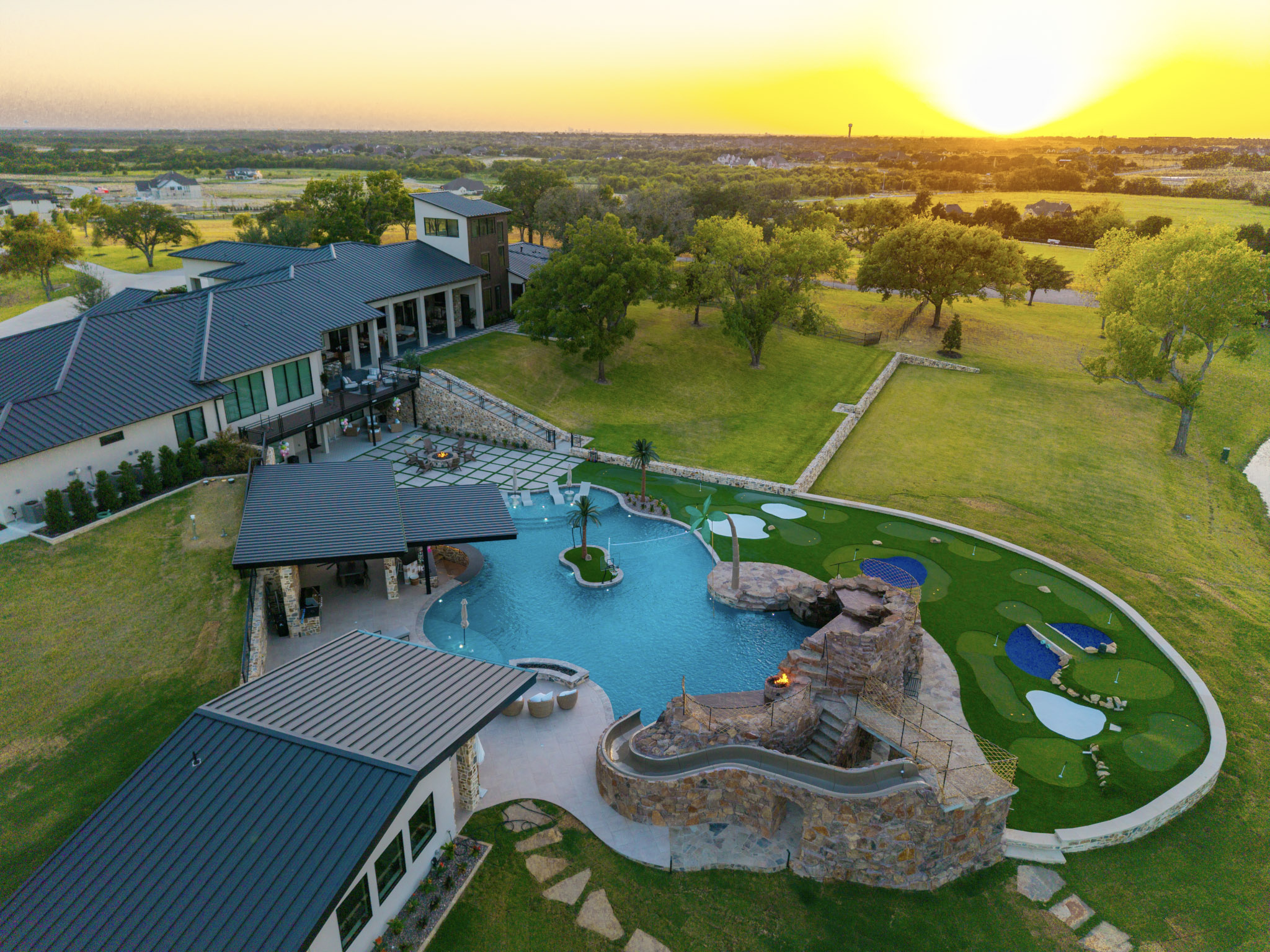 Drone view of resort-style pool, rock waterfall and large Rockwall property at sunset.