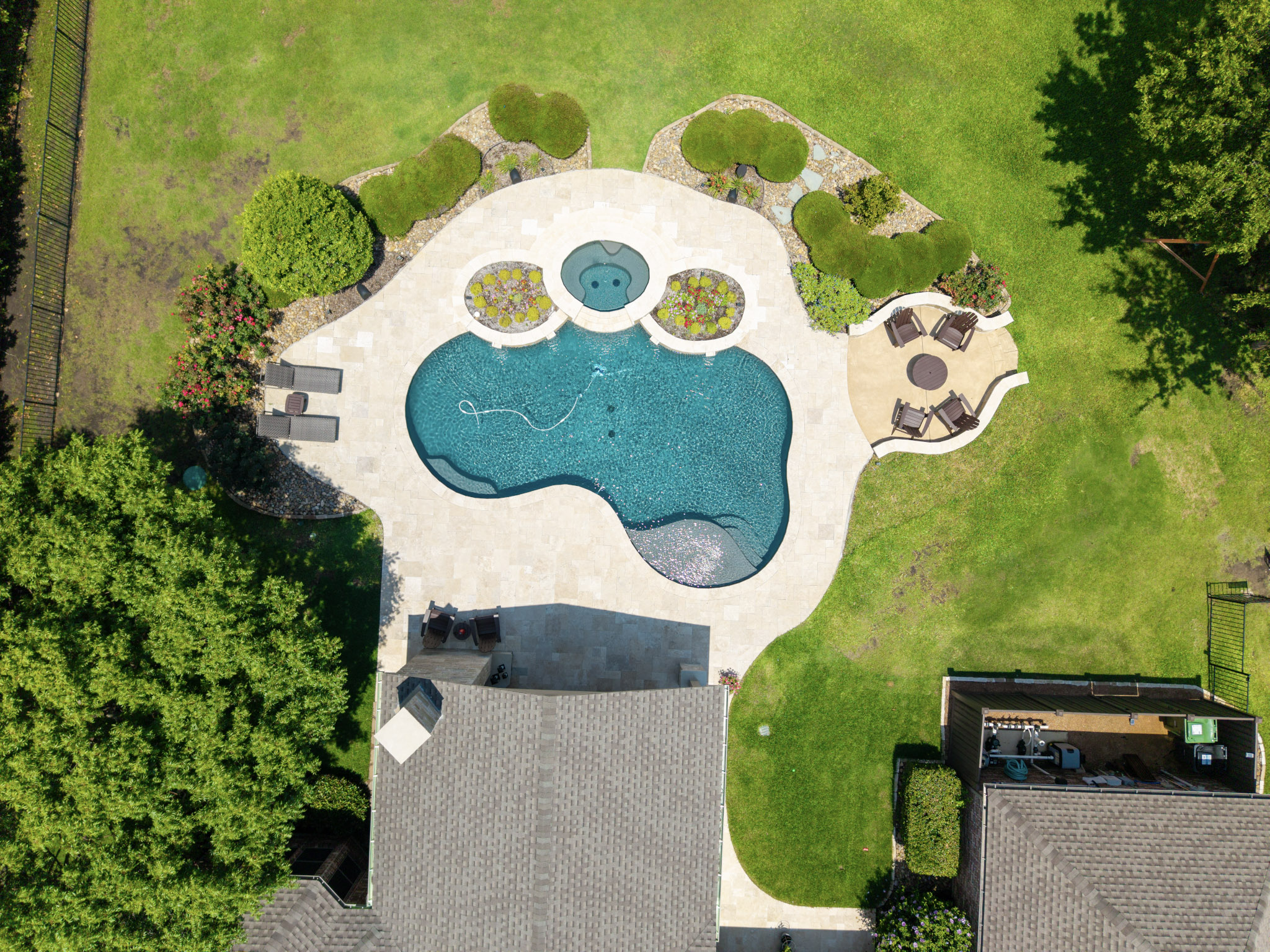 Drone view of freeform pool and spa with travertine pavers, covered patio, landscaping, and backyard seating.