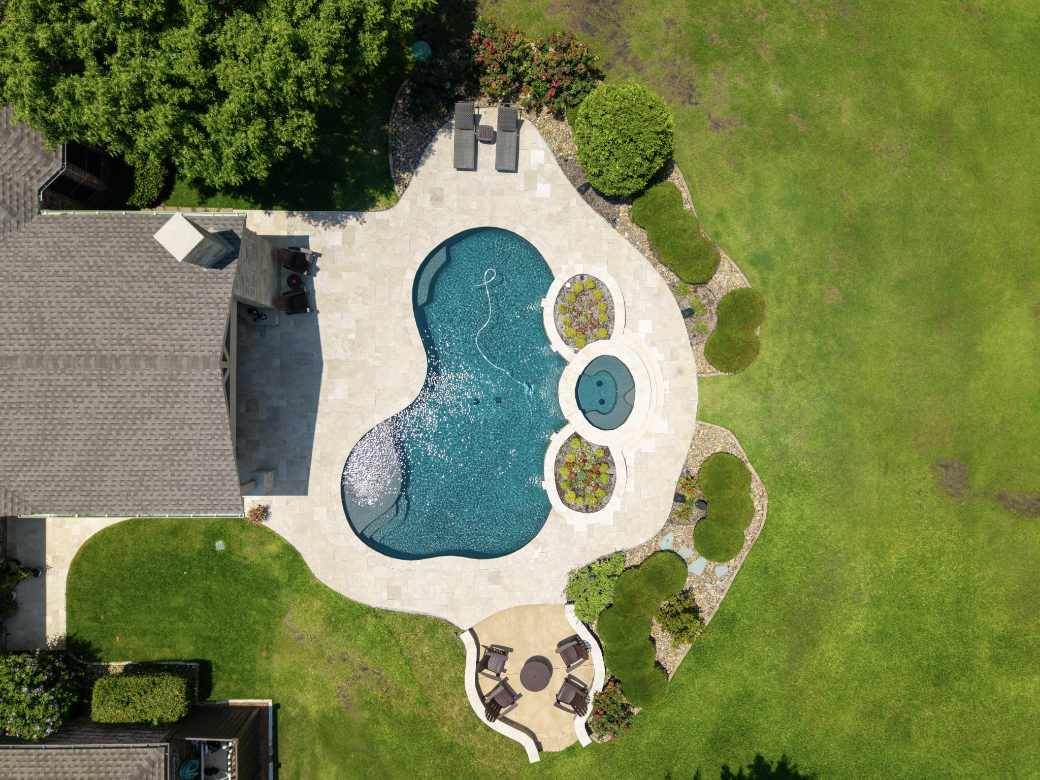 Overhead aerial view of a backyard pool, raised spa, travertine deck, patio cover, landscaping, and seating areas.