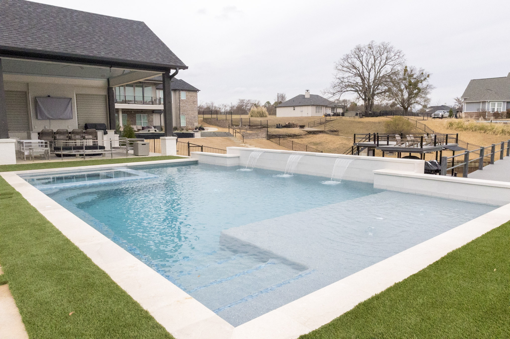 Geometric pool with large tanning ledge and three sheer descents overlooking a waterfront property.