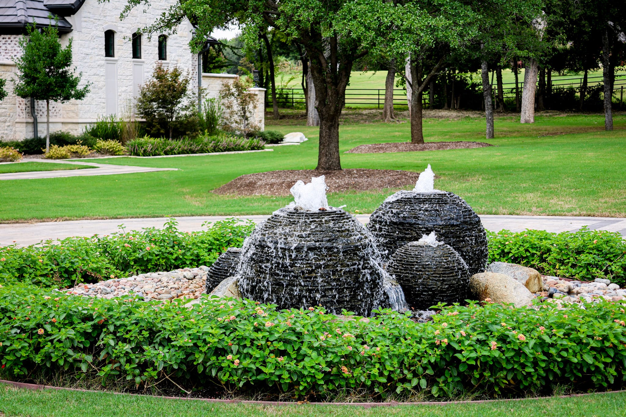 Four dark spherical fountains surrounded by decorative stone, flowering shrubs and lawn.