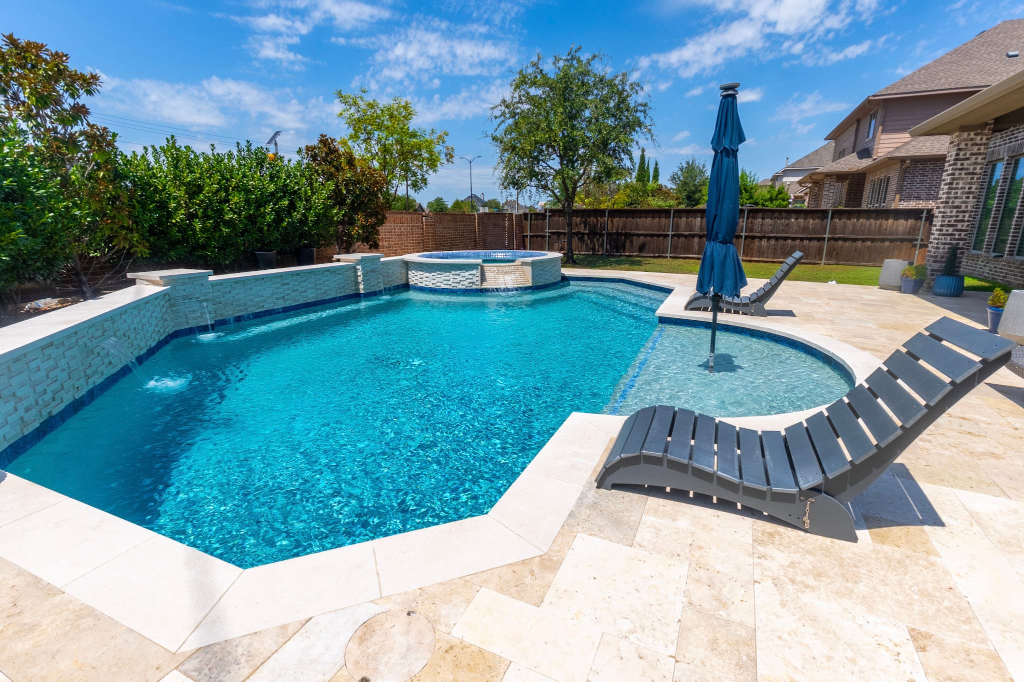 Wide view of a remodeled Frisco pool with curved sun shelf, raised spa and light travertine deck.