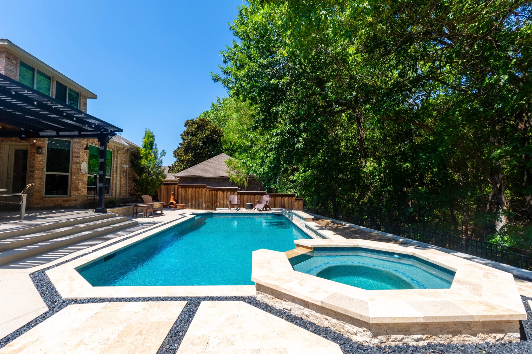 Wide view of a geometric pool and raised spa with pergola and mature trees behind the backyard.