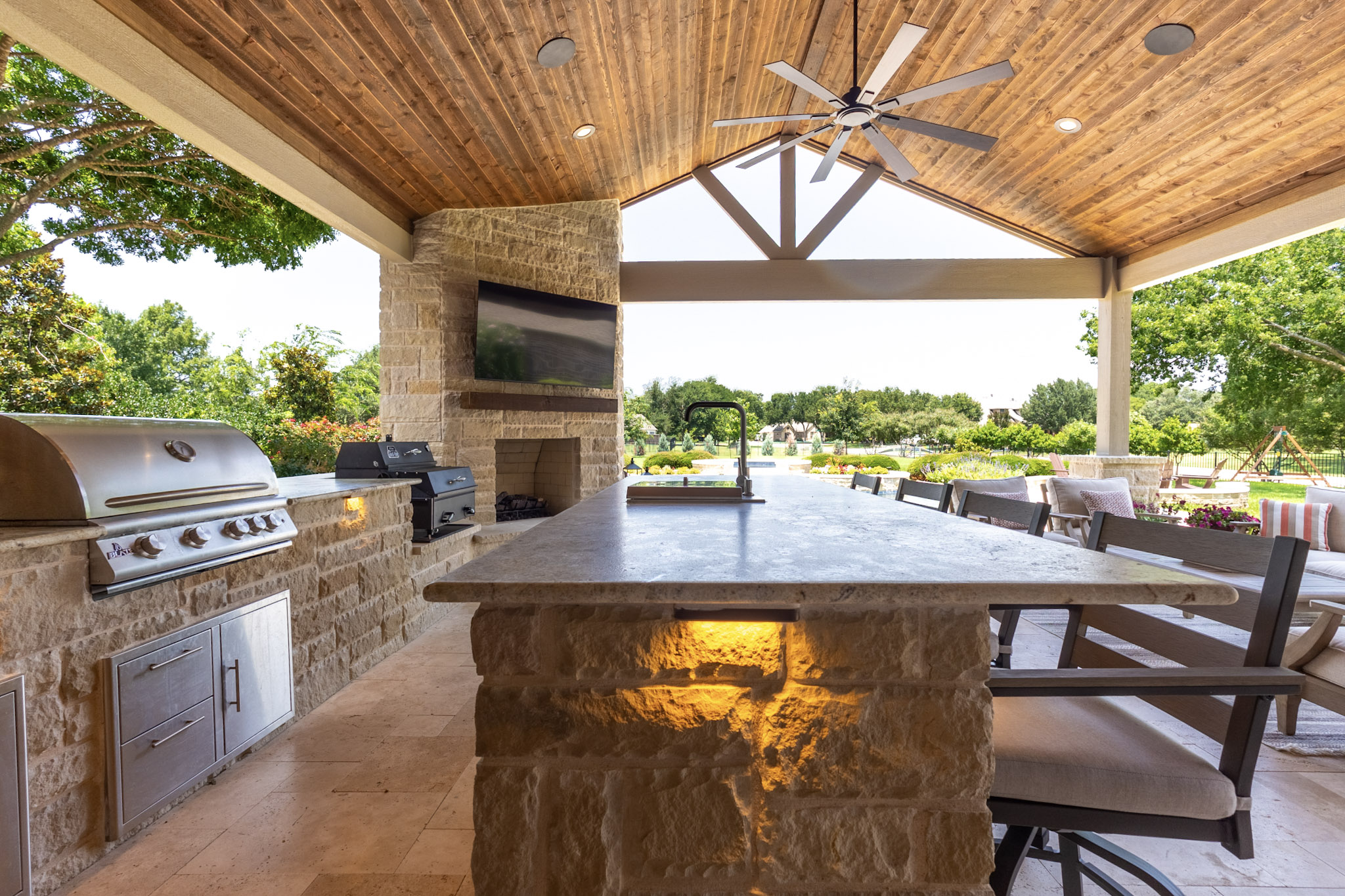 Large stone outdoor kitchen island beneath a vaulted wood patio cover with grill, sink, fireplace, and bar seating.
