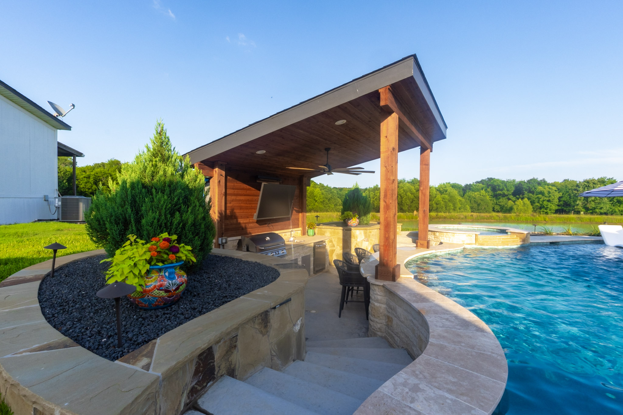 Sunken outdoor kitchen with stone walls, wood posts, built-in grill and covered patio beside a pool.