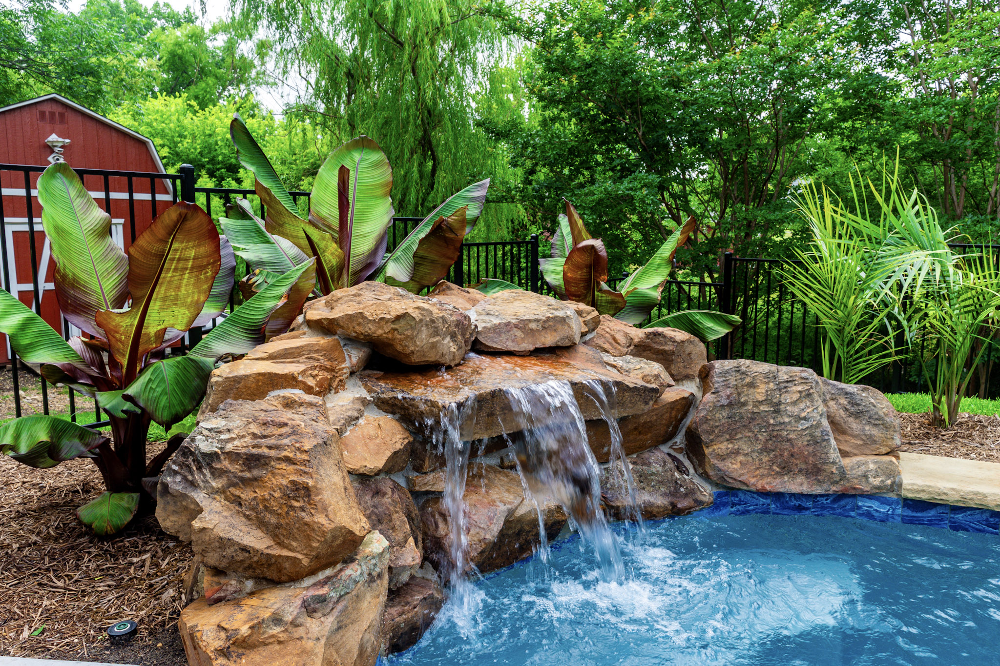 Natural rock waterfall flowing into a blue pool with tropical plants behind it.