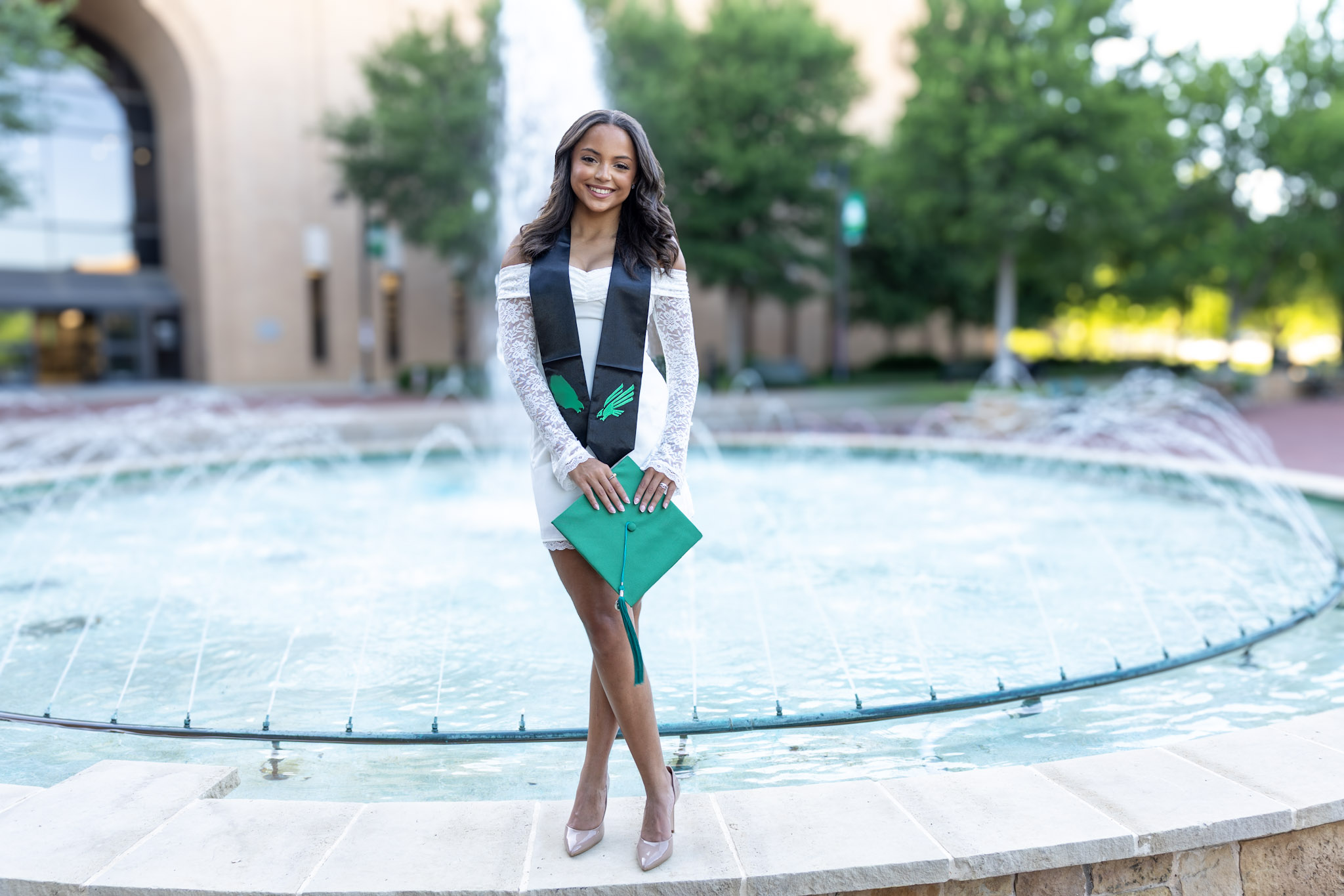 UNT graduate holding a green cap in front of a campus fountain