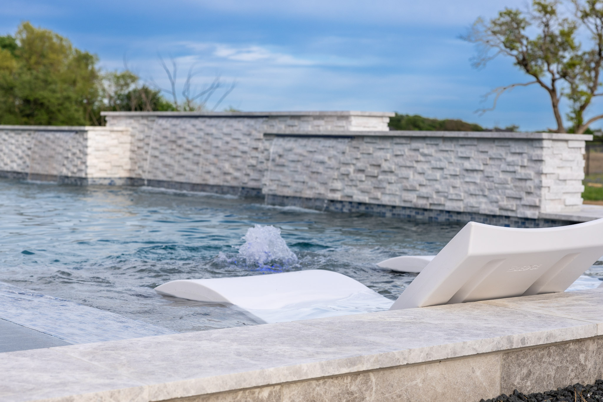 White submerged chaise lounge beside a pool bubbler and stone feature wall.