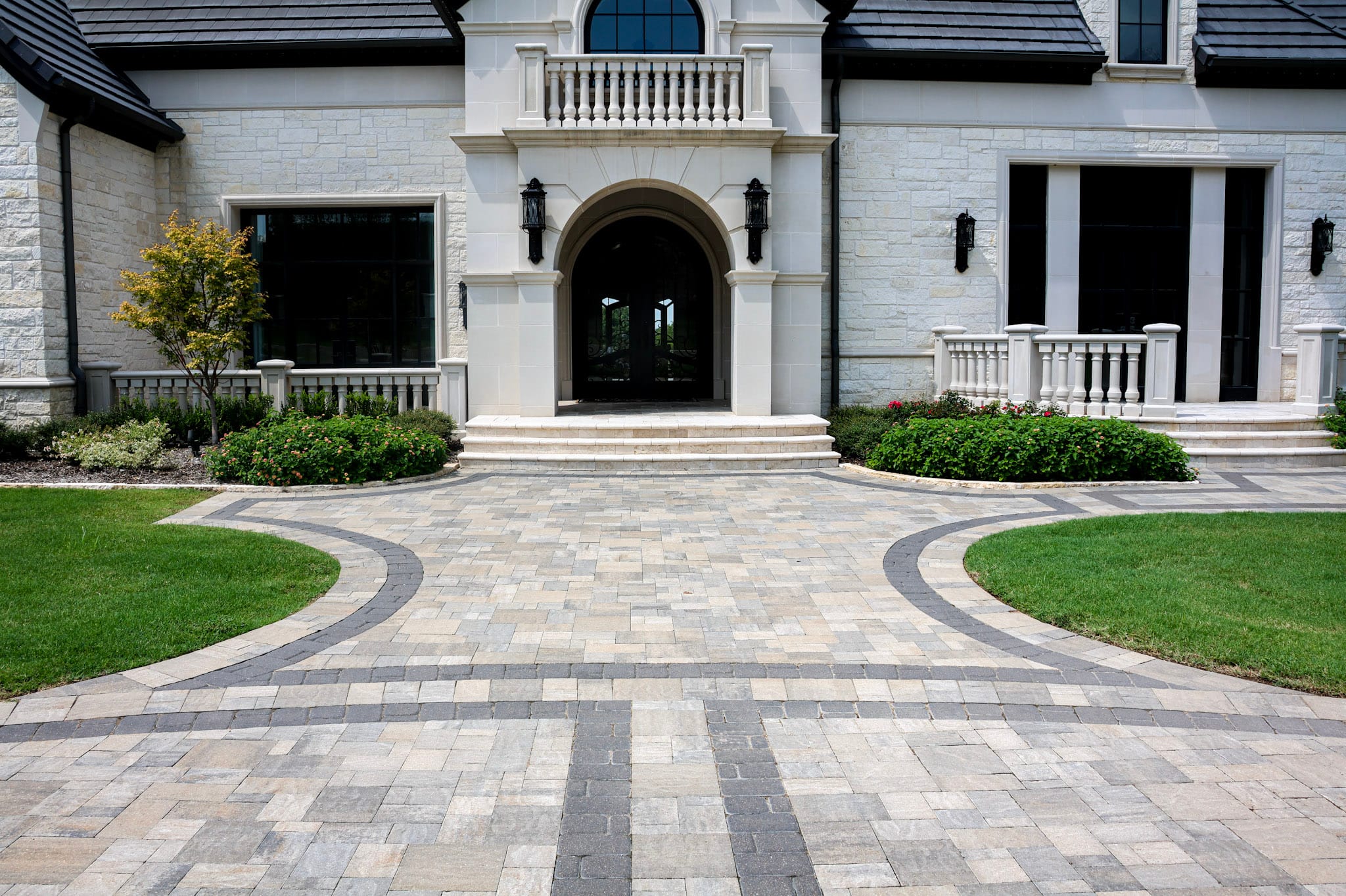 Curved Belgard paver driveway leading toward a decorative metal gate between stone columns.