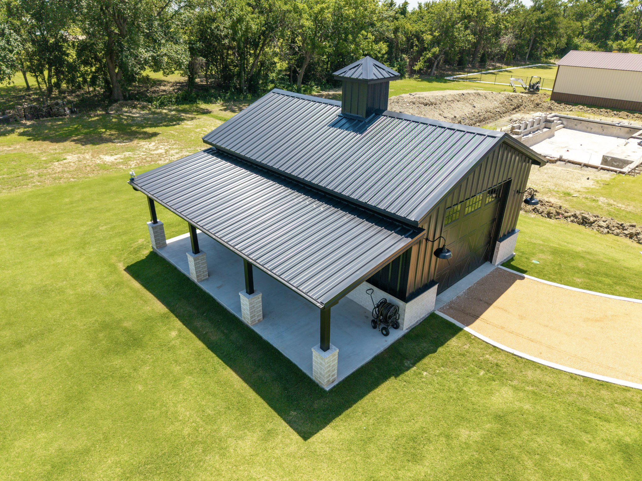 Aerial view of dark metal barn with raised roof, cupola, covered side patio and masonry columns.