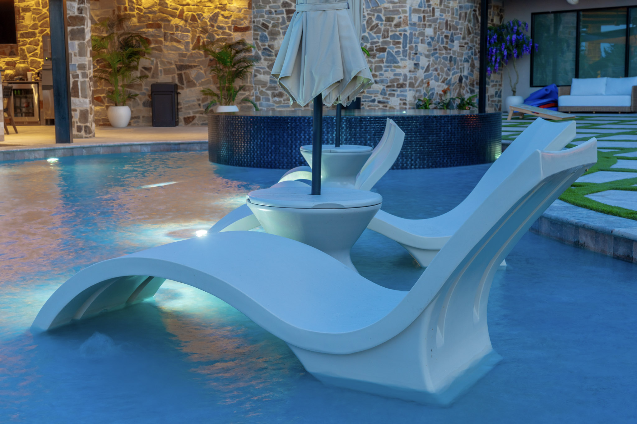 Submerged white pool loungers and umbrella table illuminated at dusk.