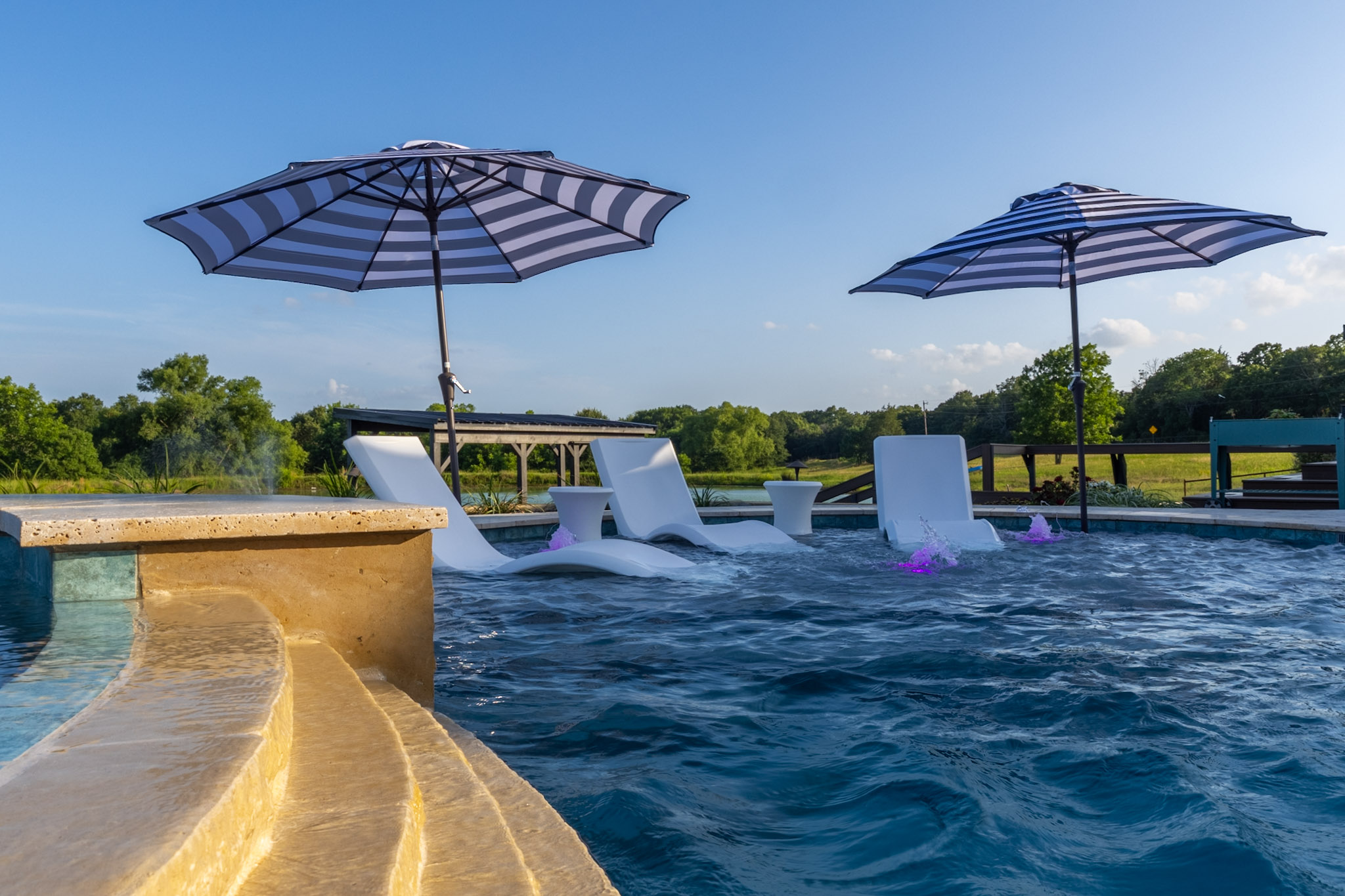 Pool-level view of a tanning ledge with white loungers, striped umbrellas and purple-lit bubblers.