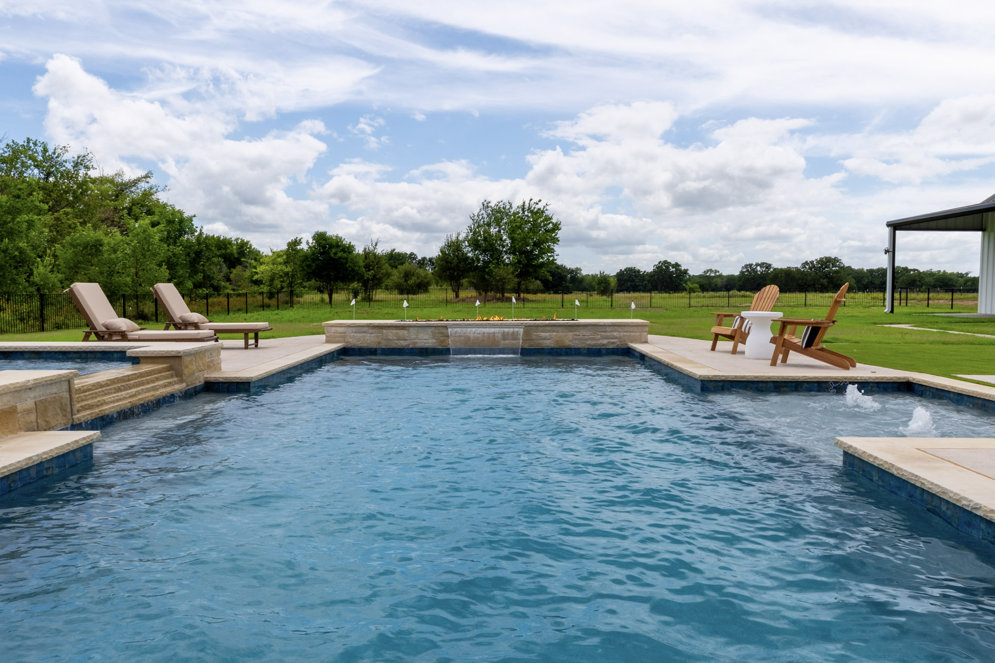 Wide view across a custom swimming pool toward a raised wall with sheer descent waterfall and linear fire feature.
