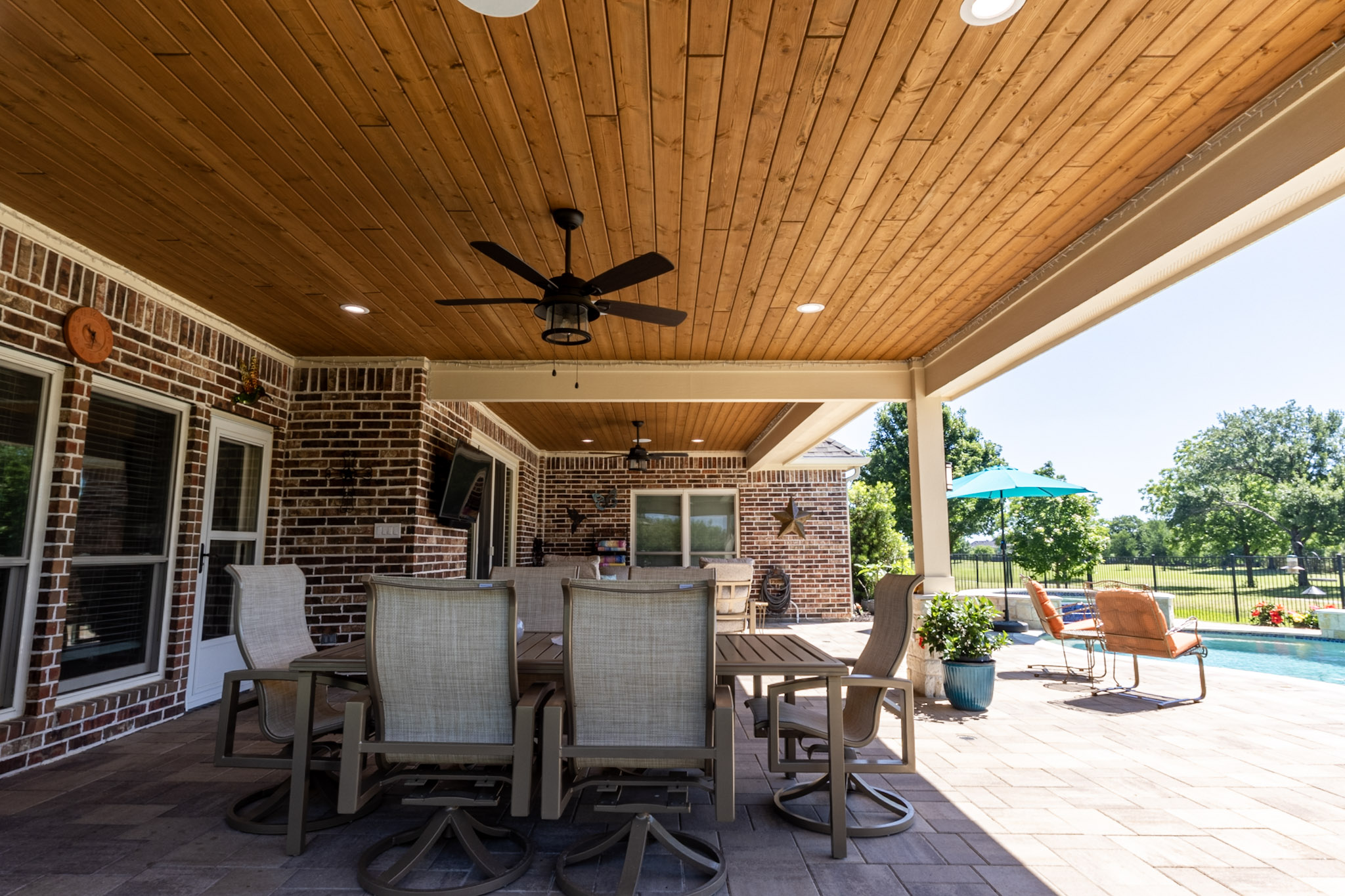 Outdoor dining table beneath a wood ceiling with ceiling fan and pool visible beside the patio.