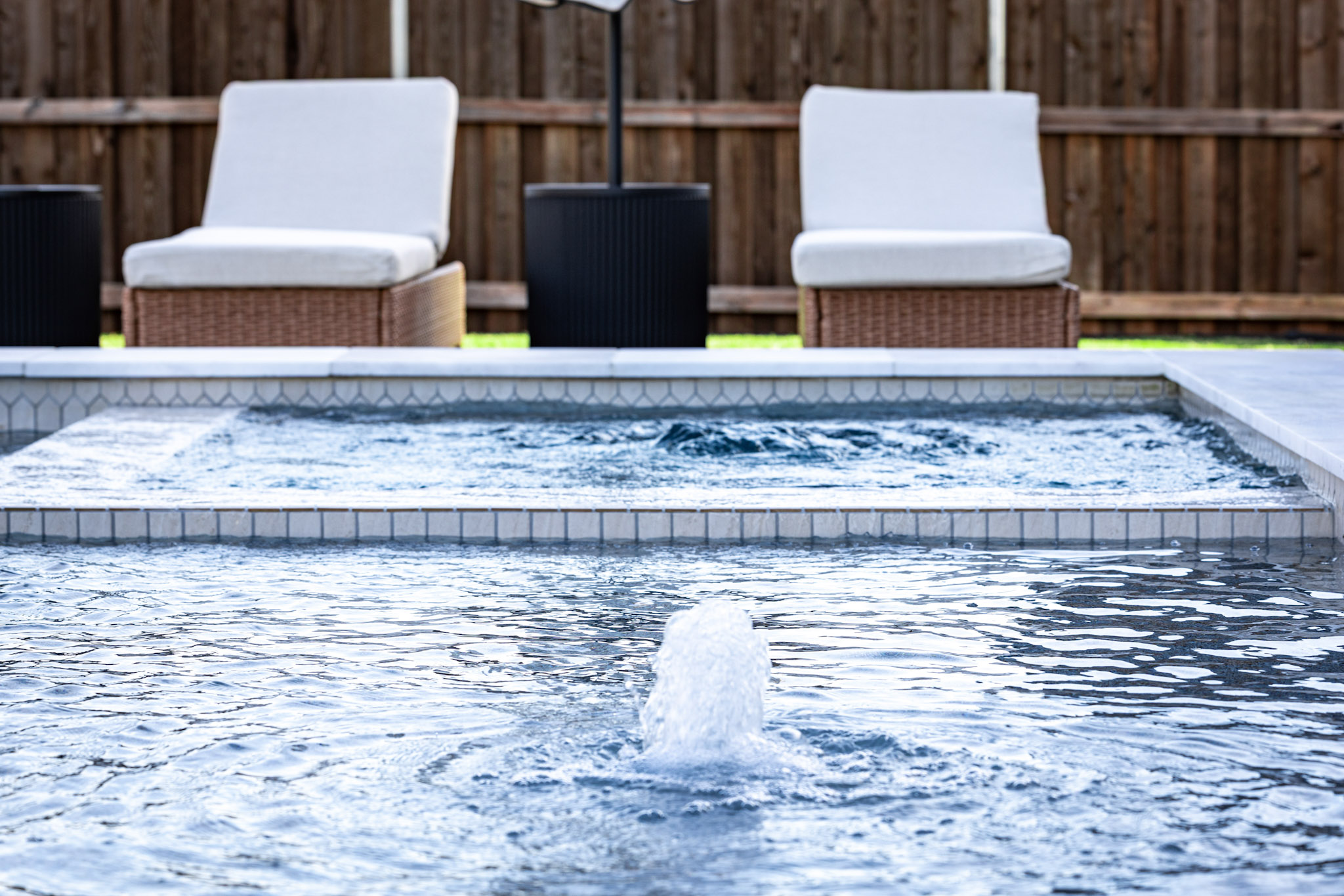 Active bubbler in pool water with square spa and two cushioned chairs behind it.