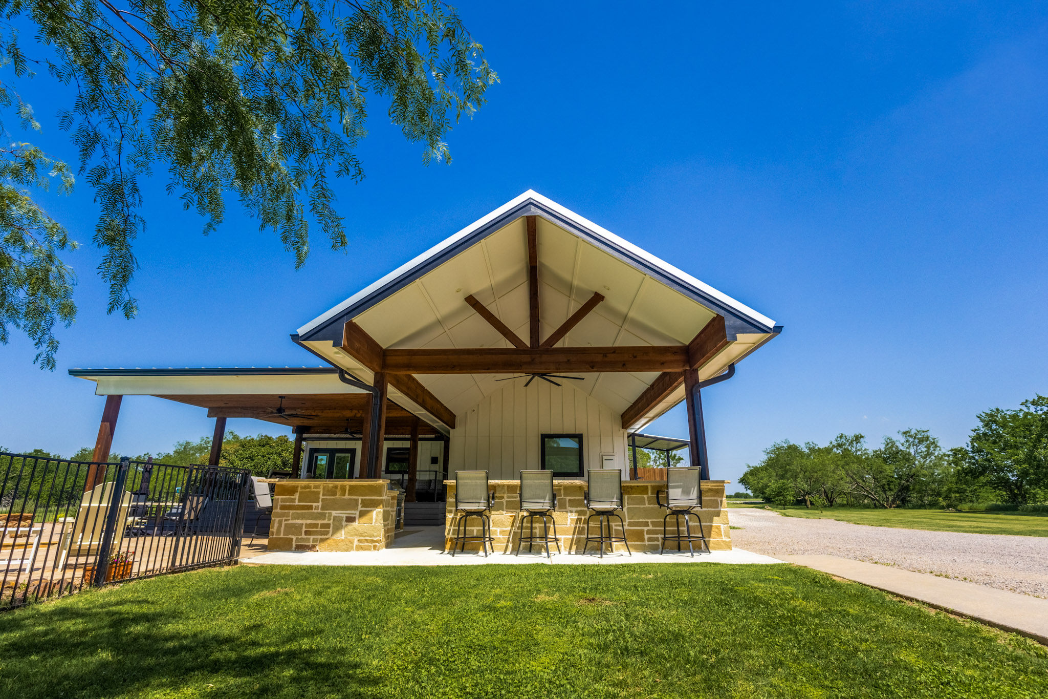 Covered stone outdoor kitchen with wood beams, grill, deck stairs and pool beyond.