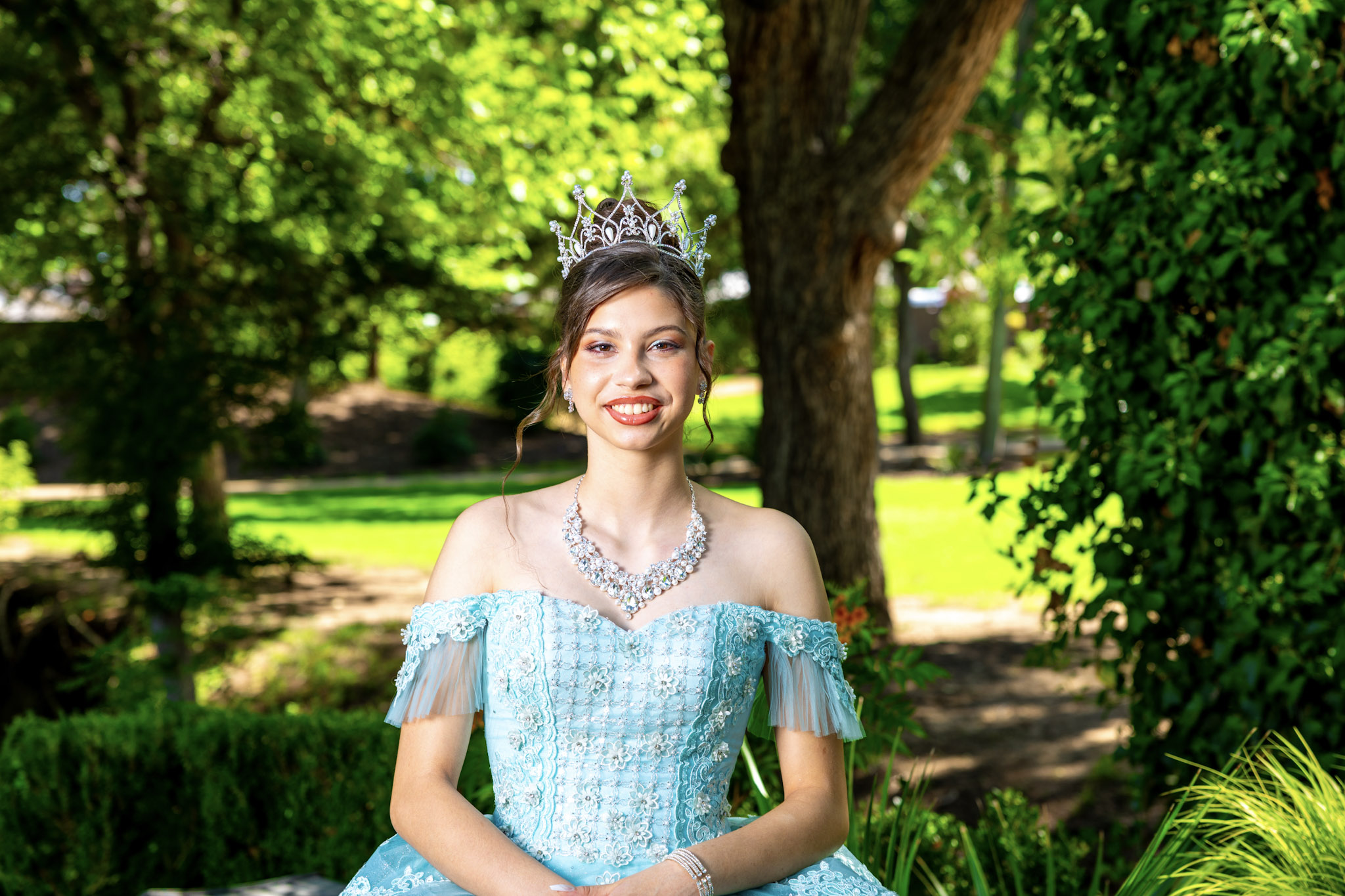 Quinceañera in aqua gown and crown posing beneath mature trees