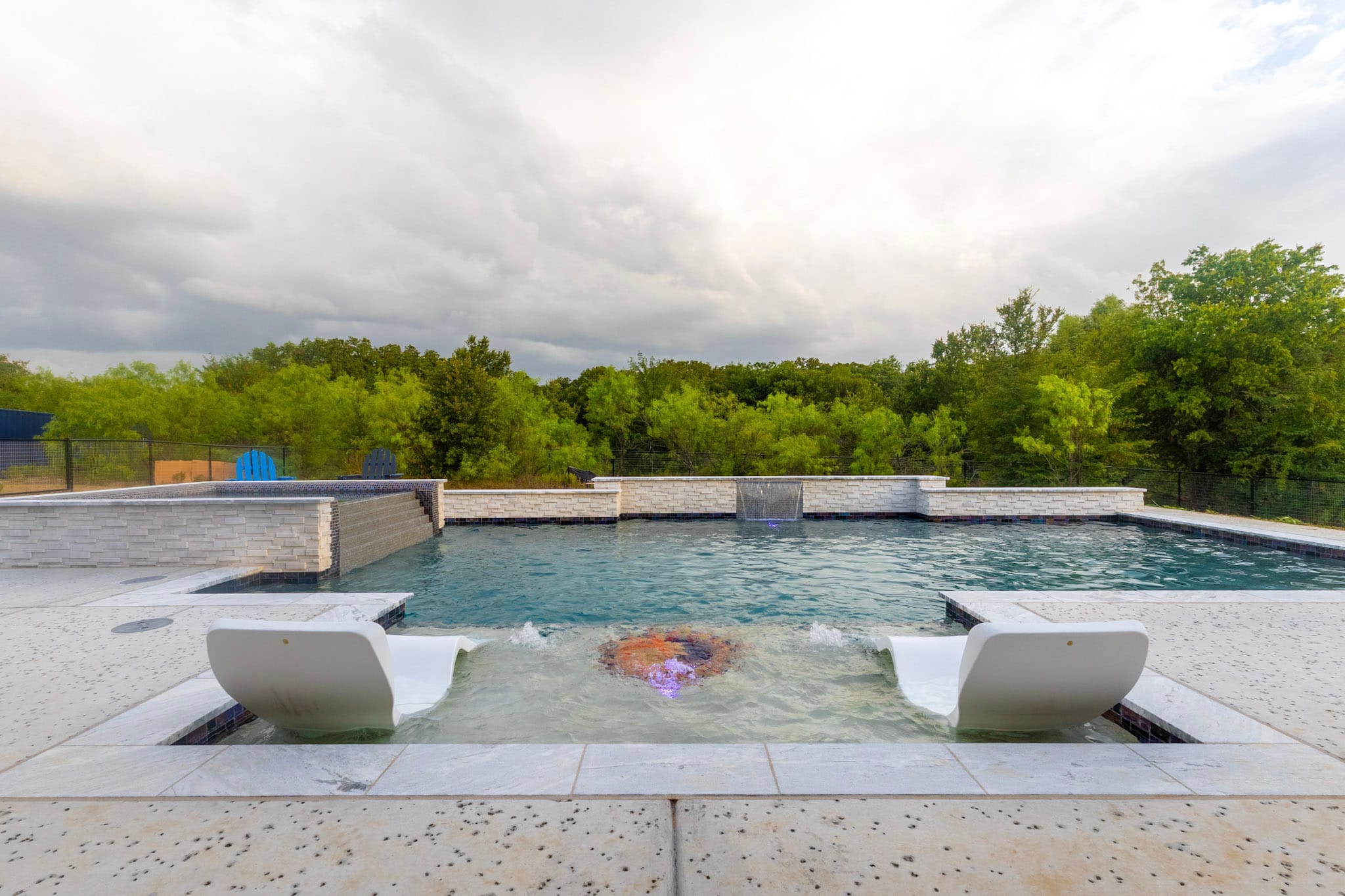 Tanning ledge with two in-pool loungers and a bubbler overlooking a custom pool with a raised stone water-feature wall.
