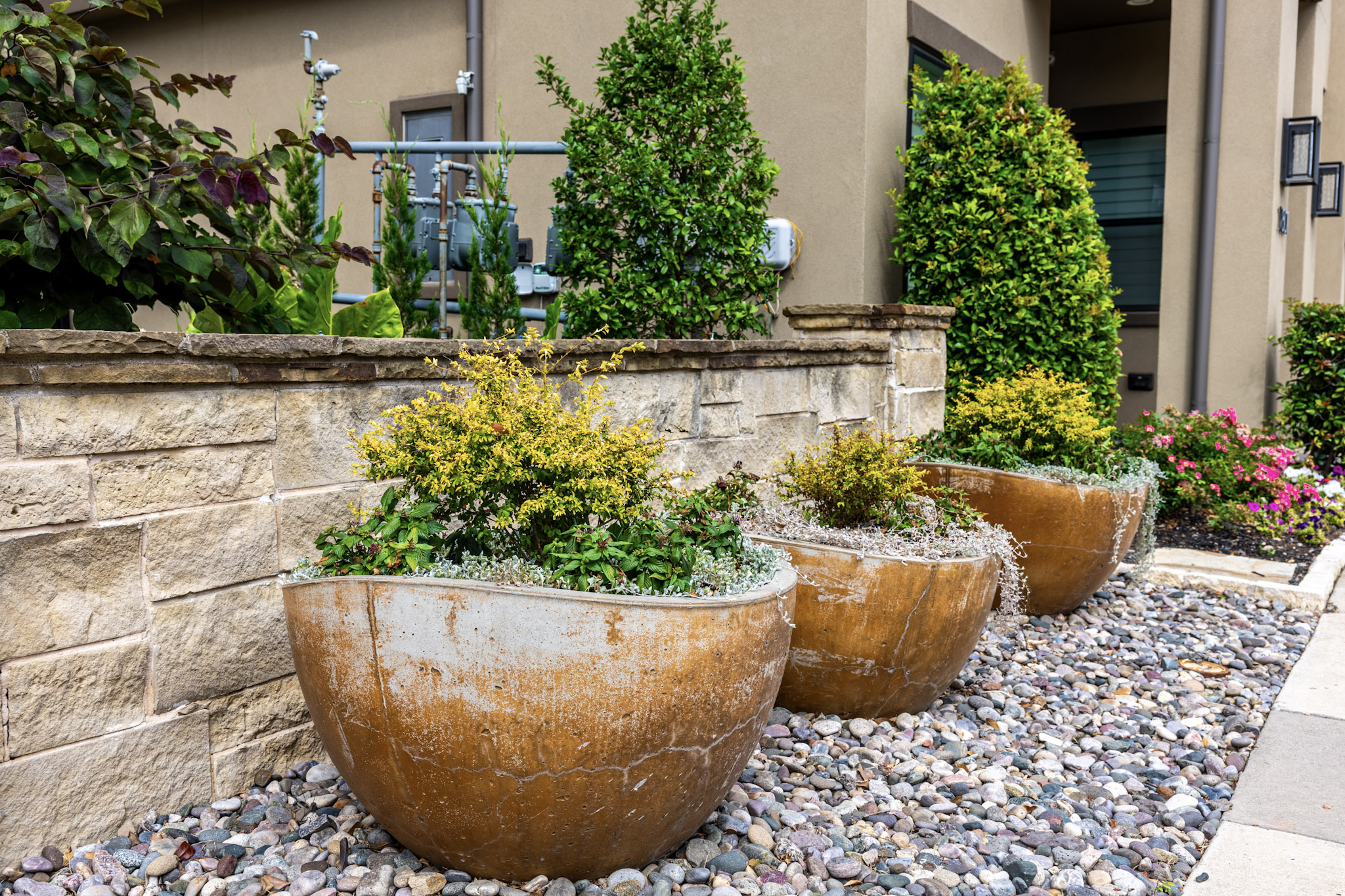 Three brown planters with shrubs beside a stone wall and river rock.