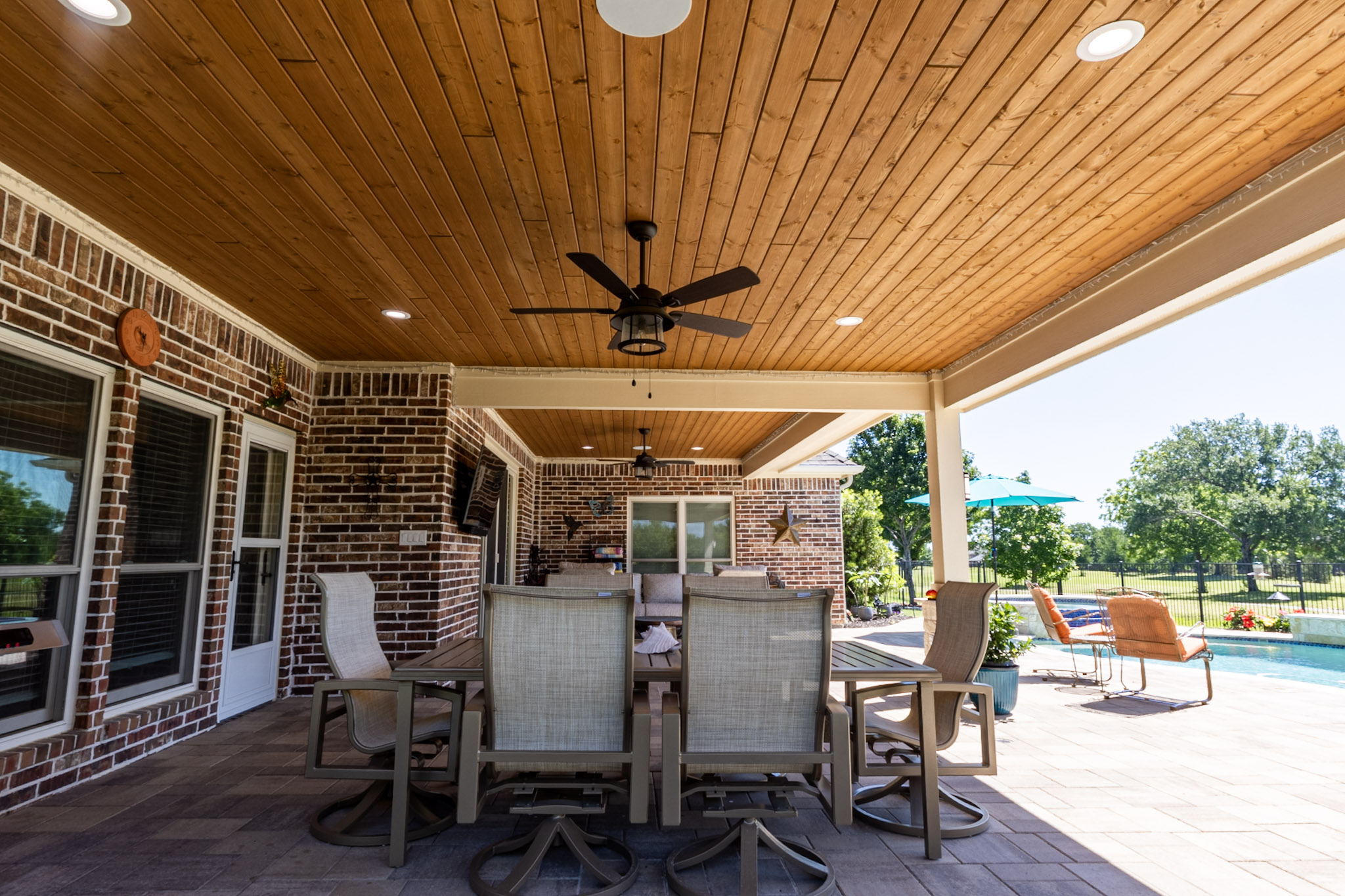 Covered patio dining table with wood ceiling and swimming pool visible beyond.