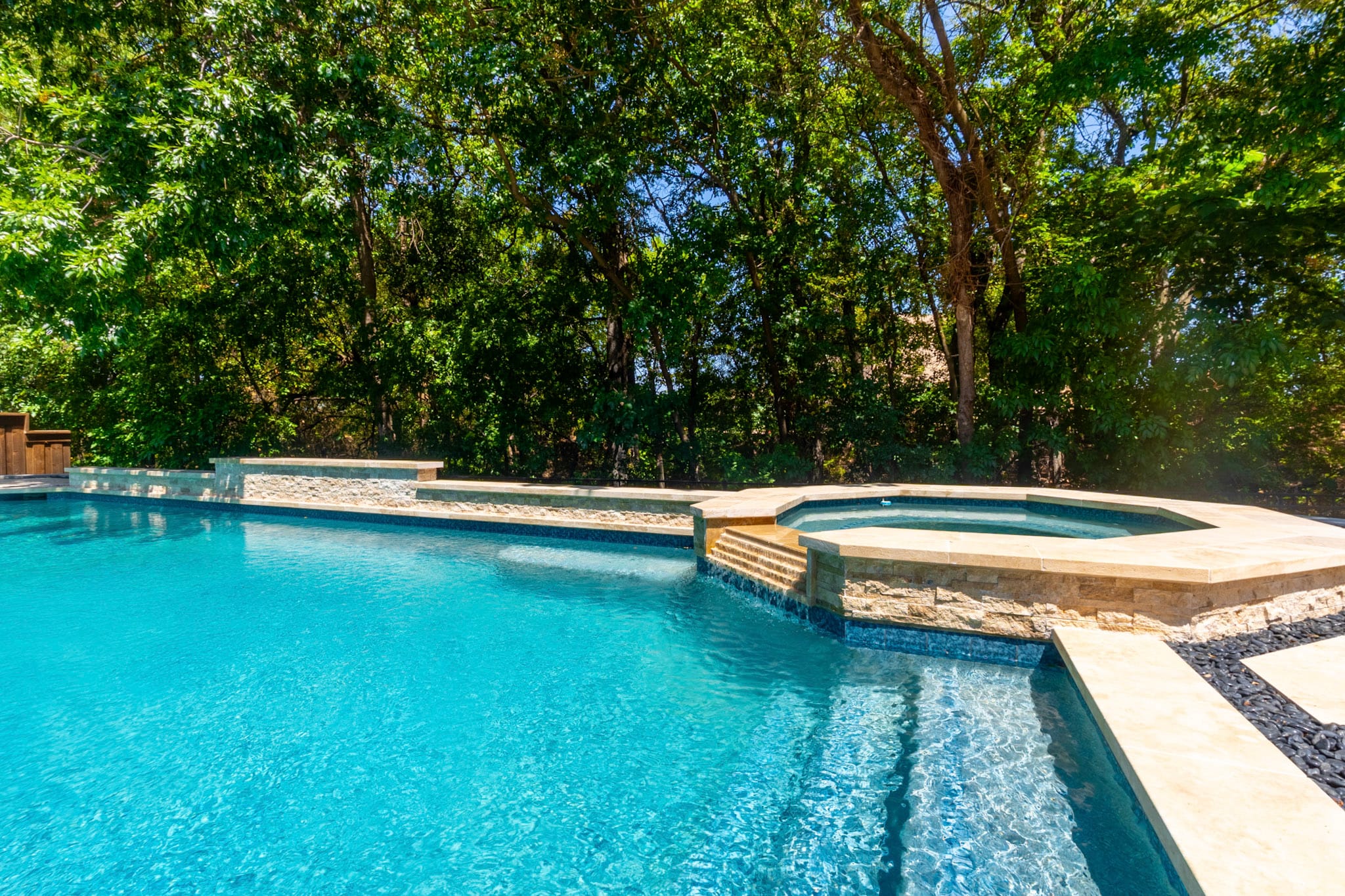 Wide pool view with raised spa, tiered spillover and stone water feature wall beneath mature trees.