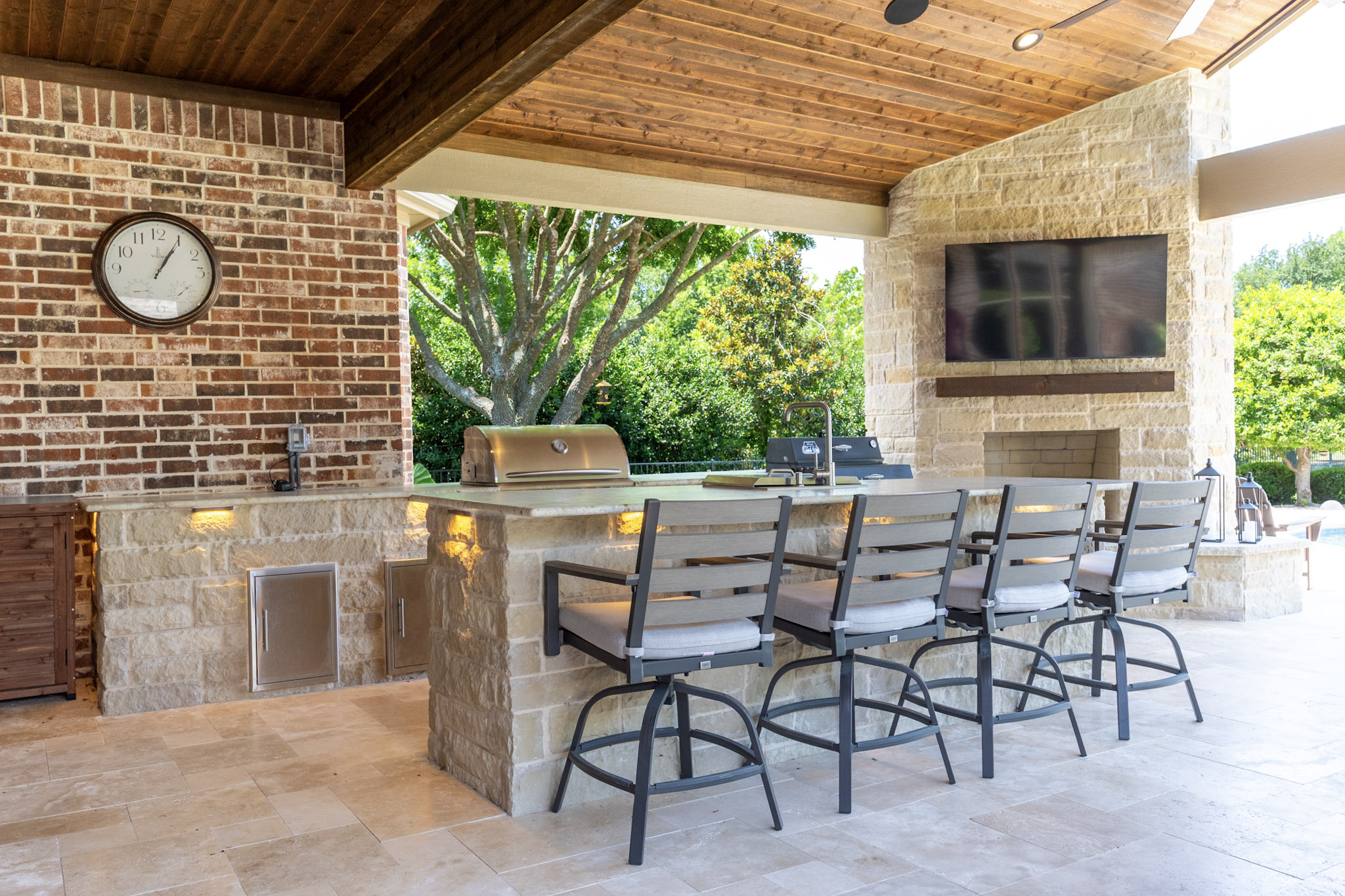 Outdoor kitchen bar with stone masonry, bar stools, built-in grill, fireplace, television, and wood ceiling.