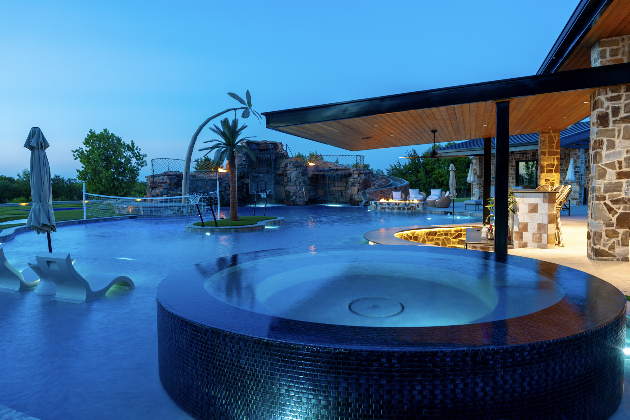 Circular glass-tile spa overlooking illuminated resort-style swimming pool.