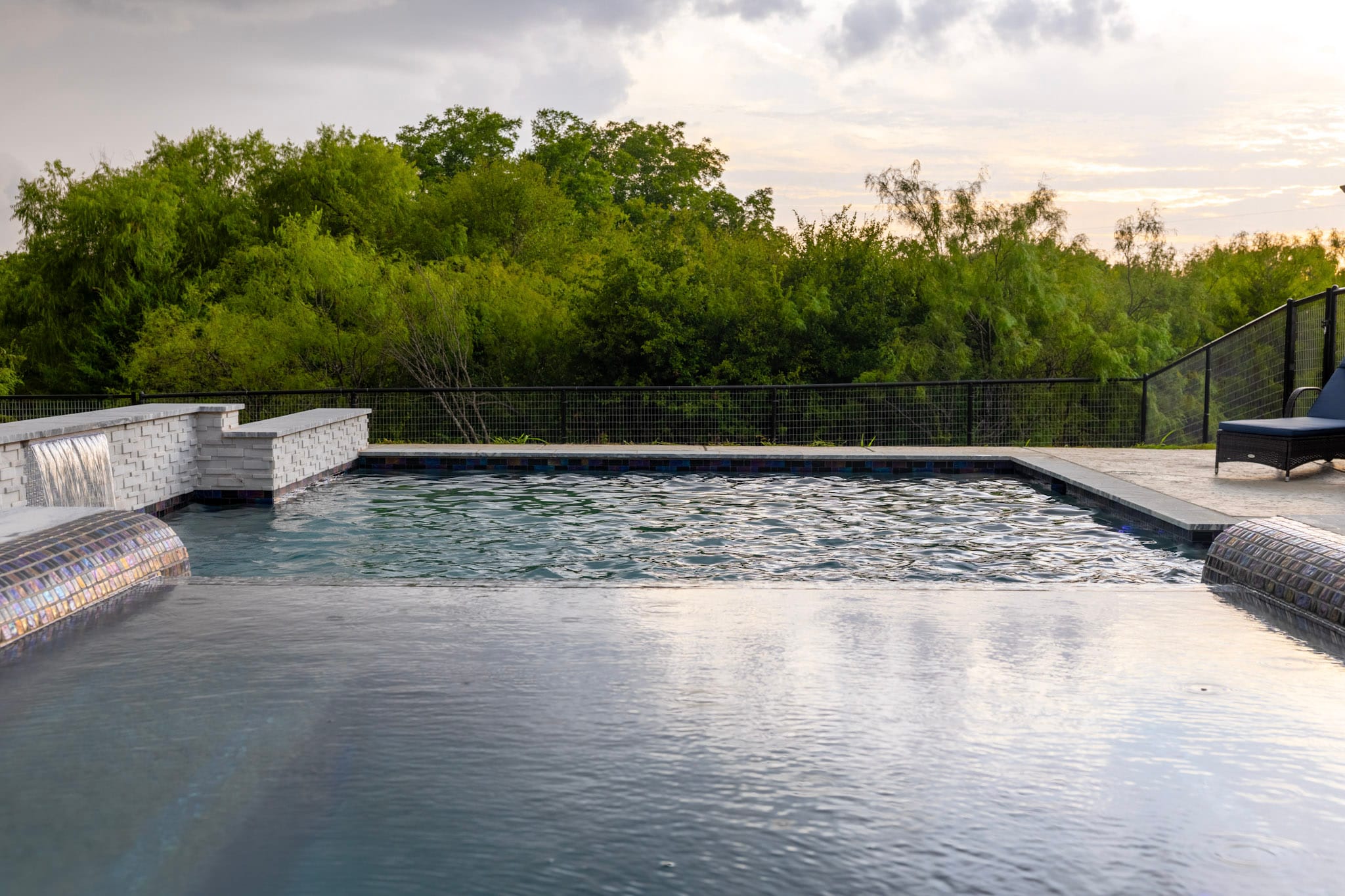 Low view across a raised spa toward a large custom pool and wooded North Texas landscape.