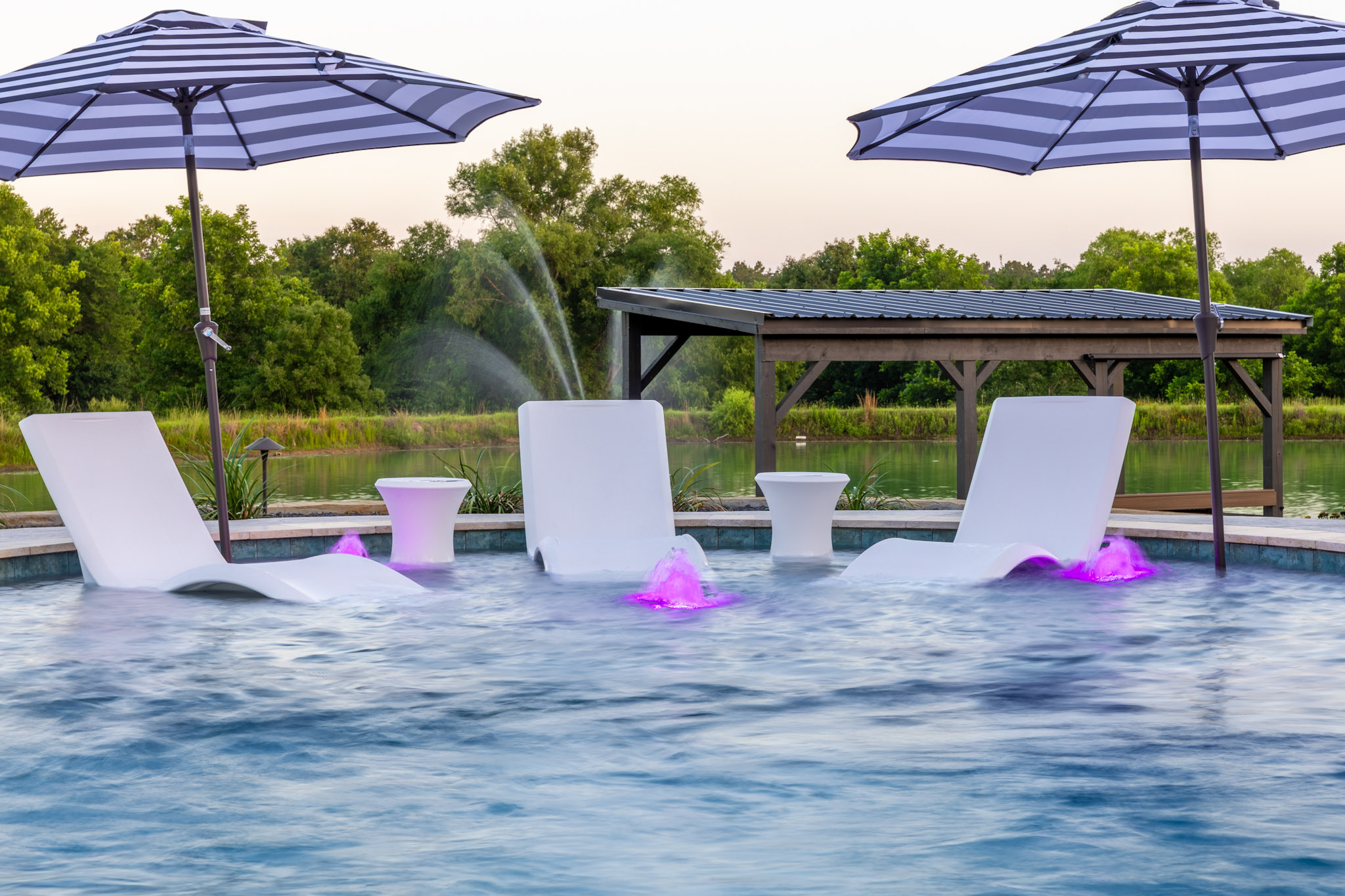 Three white in-pool loungers with striped umbrellas and purple-lit bubblers on a tanning ledge.