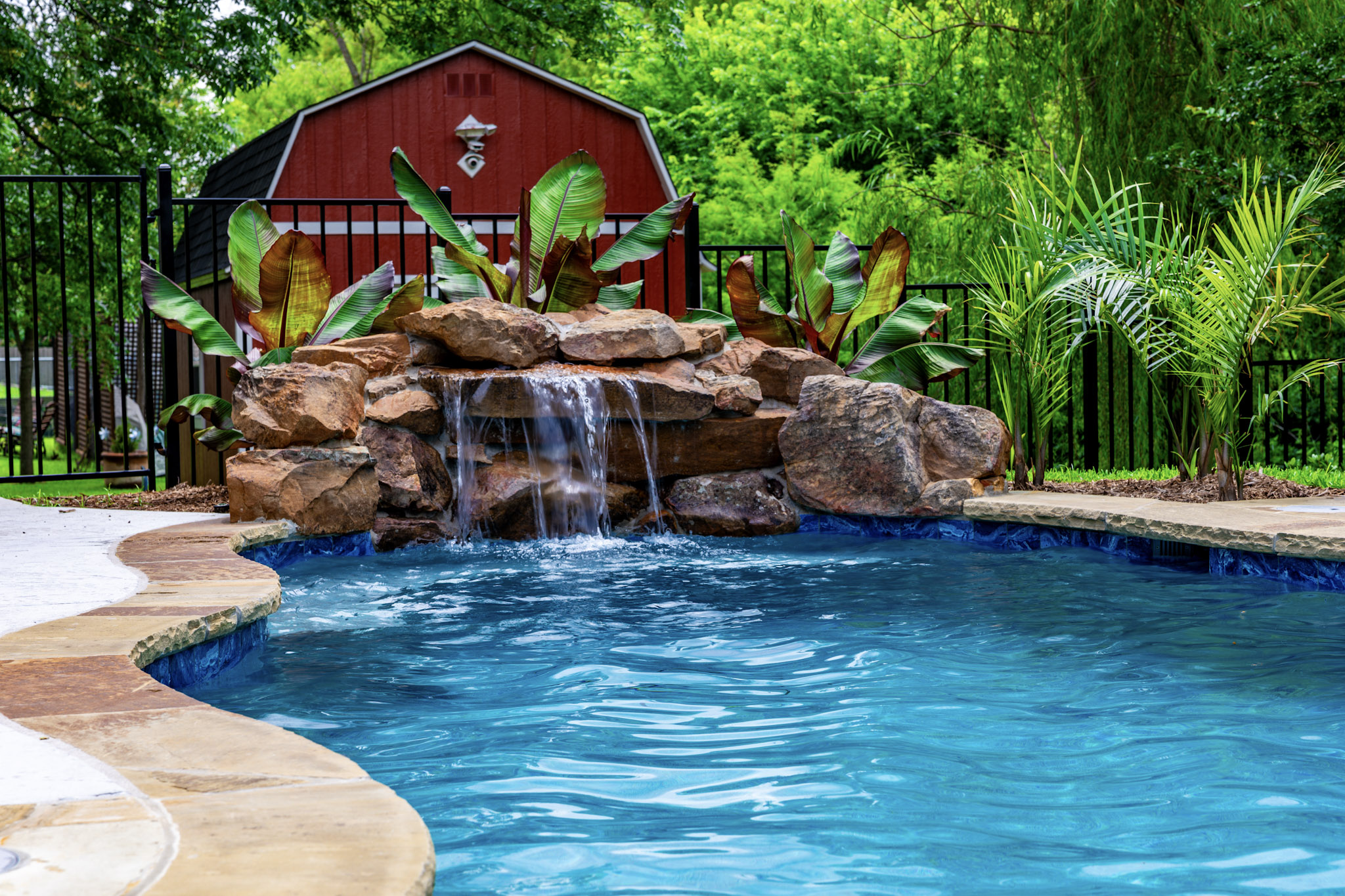 Swimming pool facing a natural rock waterfall surrounded by tropical plants.