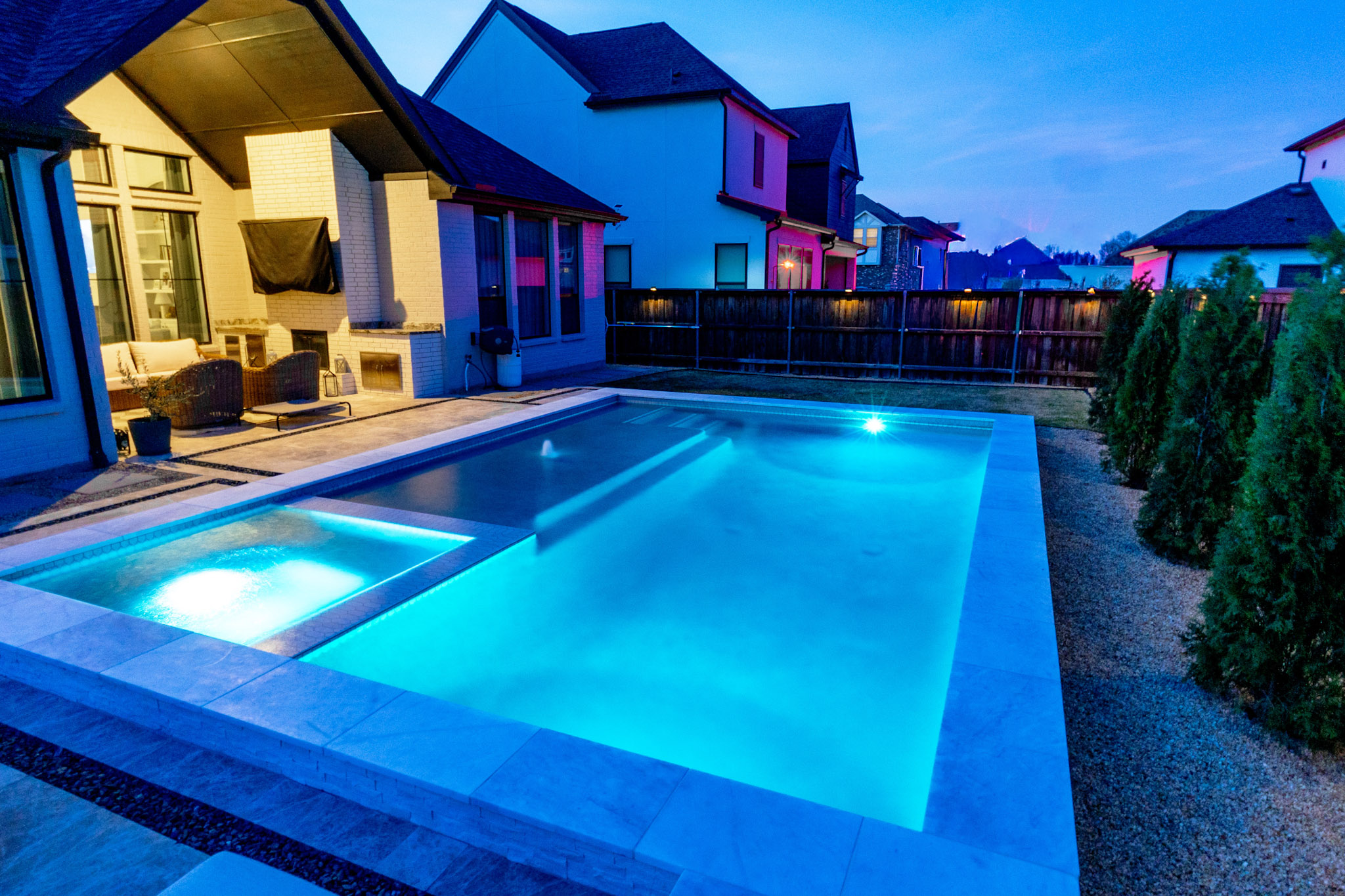 Illuminated pool and square spa beside a covered patio and white brick home at dusk.
