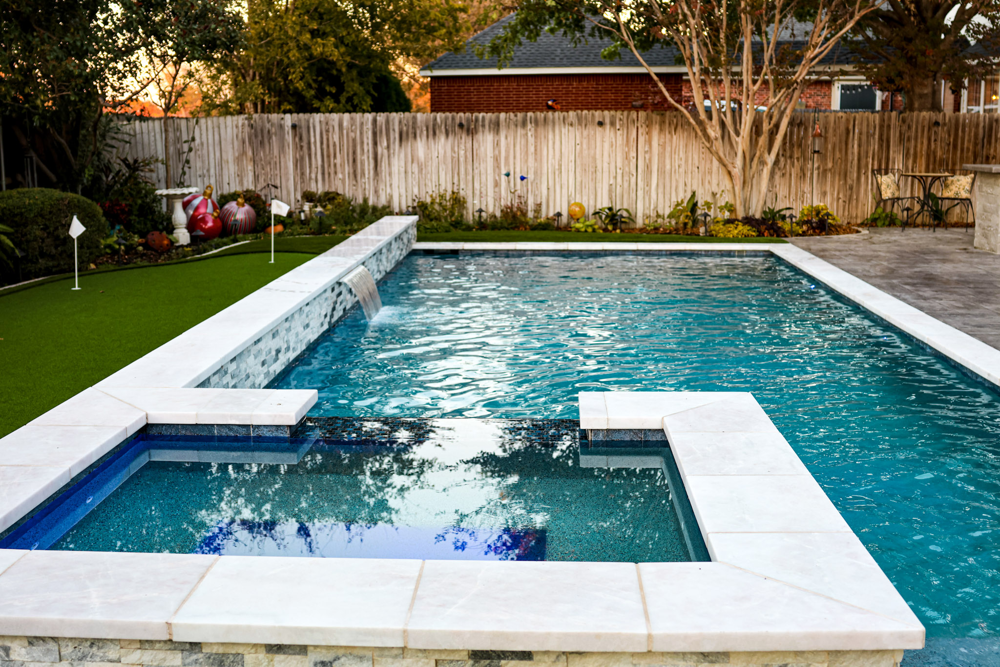 Raised square spa beside a blue pool with a covered fireplace patio in the background.