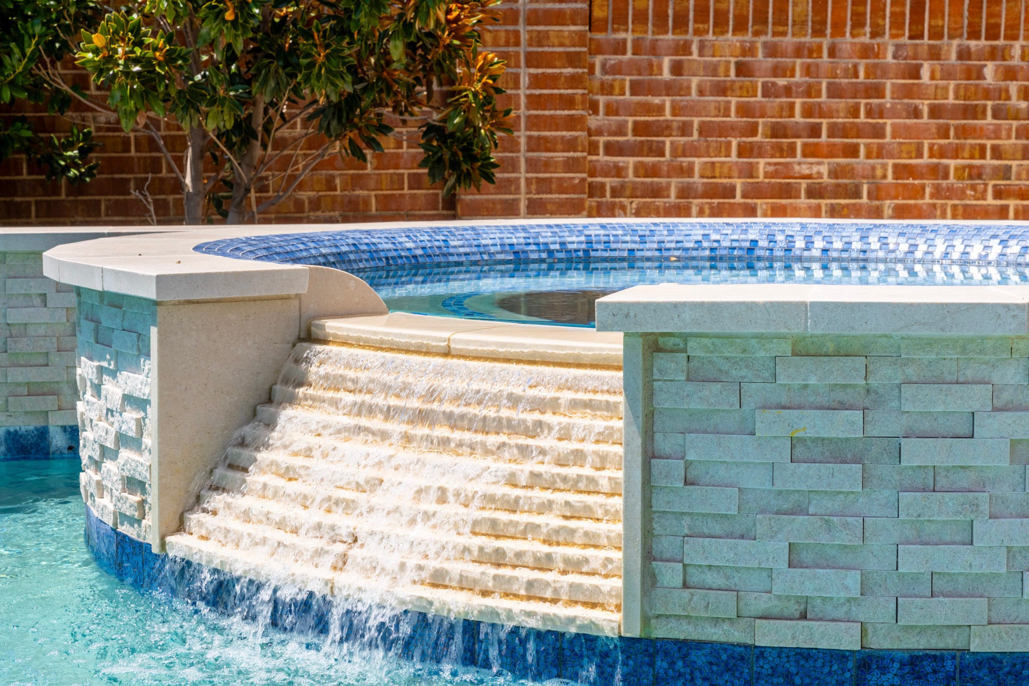 Close-up of a stepped spa waterfall flowing between light stacked-stone walls into a swimming pool.