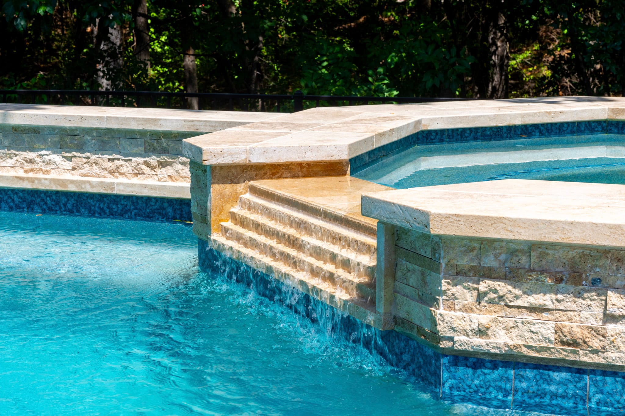 Tiered water spillover flowing from a raised stone spa into a blue swimming pool.