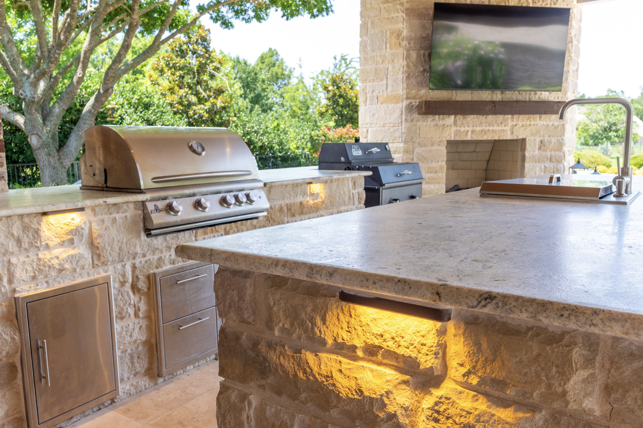 Stone outdoor kitchen island with sink, built-in grill, illuminated masonry, fireplace, and television.
