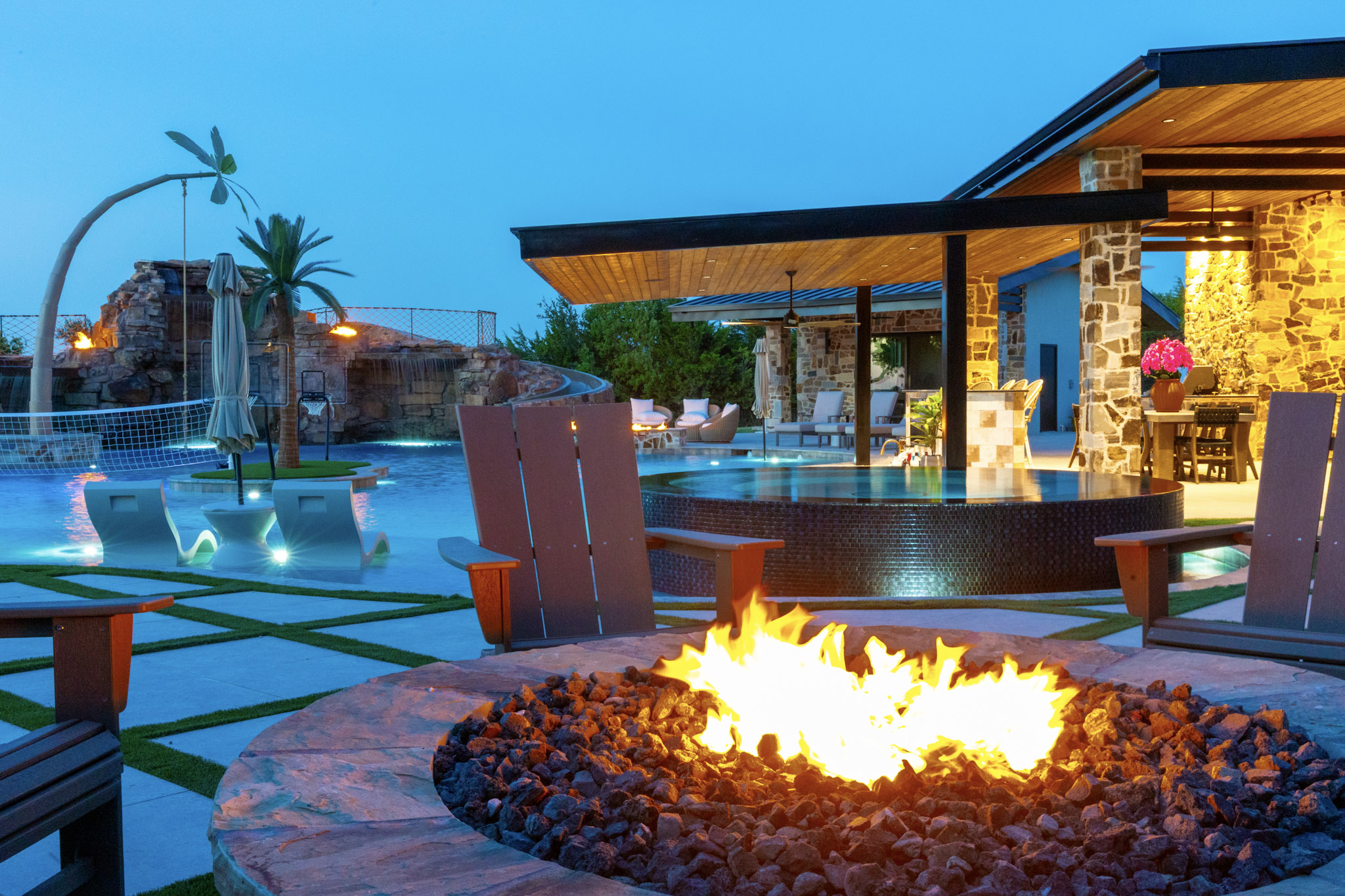 Large circular fire pit overlooking illuminated resort pool and pavilion.