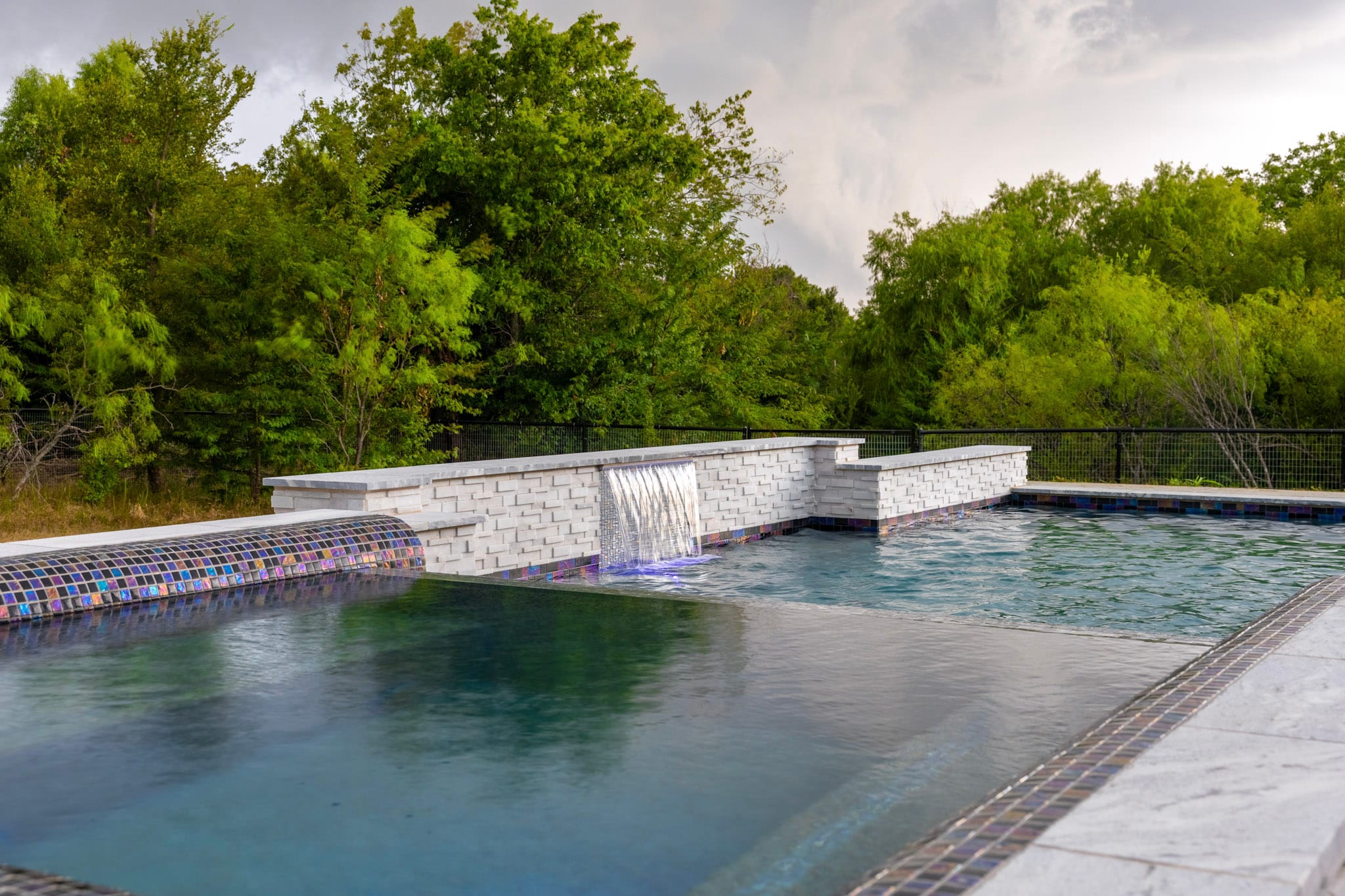 Raised white stone pool wall with a central sheer descent flowing into a custom swimming pool.