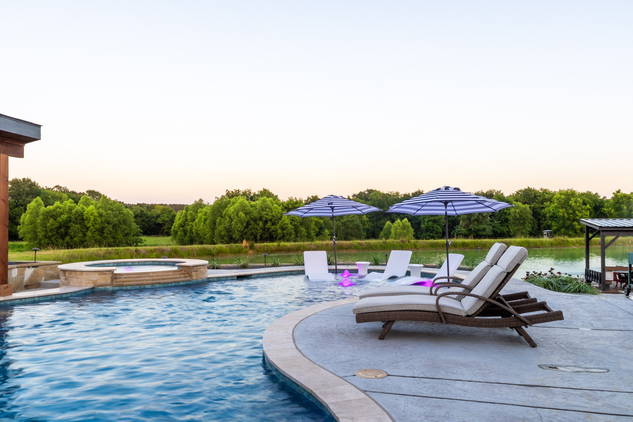 Freeform pool with poolside loungers, tanning ledge, umbrellas and raised spa beside a private lake.