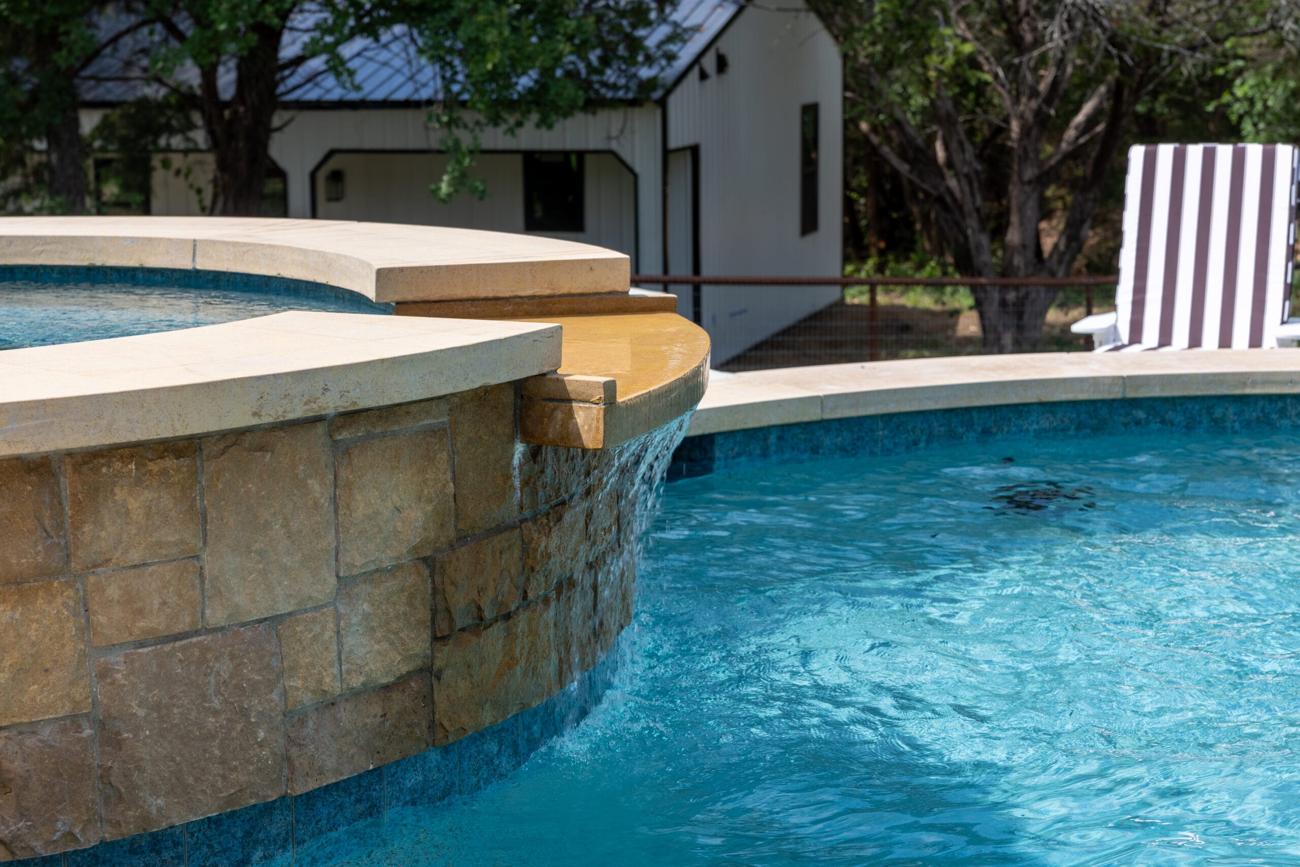 Curved stone spa with a narrow spillway flowing into blue swimming pool water.