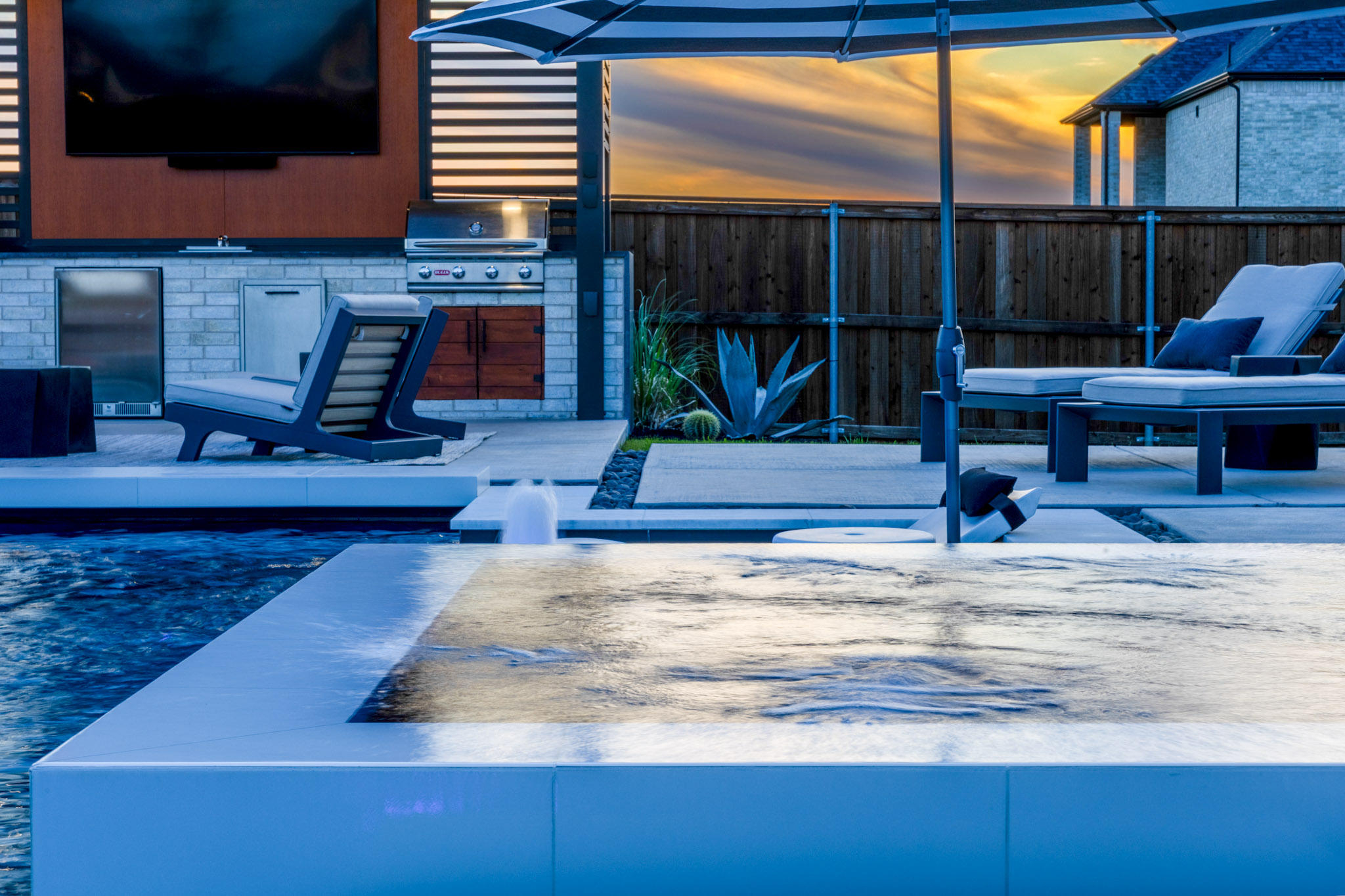Raised spa overlooking modern outdoor kitchen and poolside lounge area at sunset.