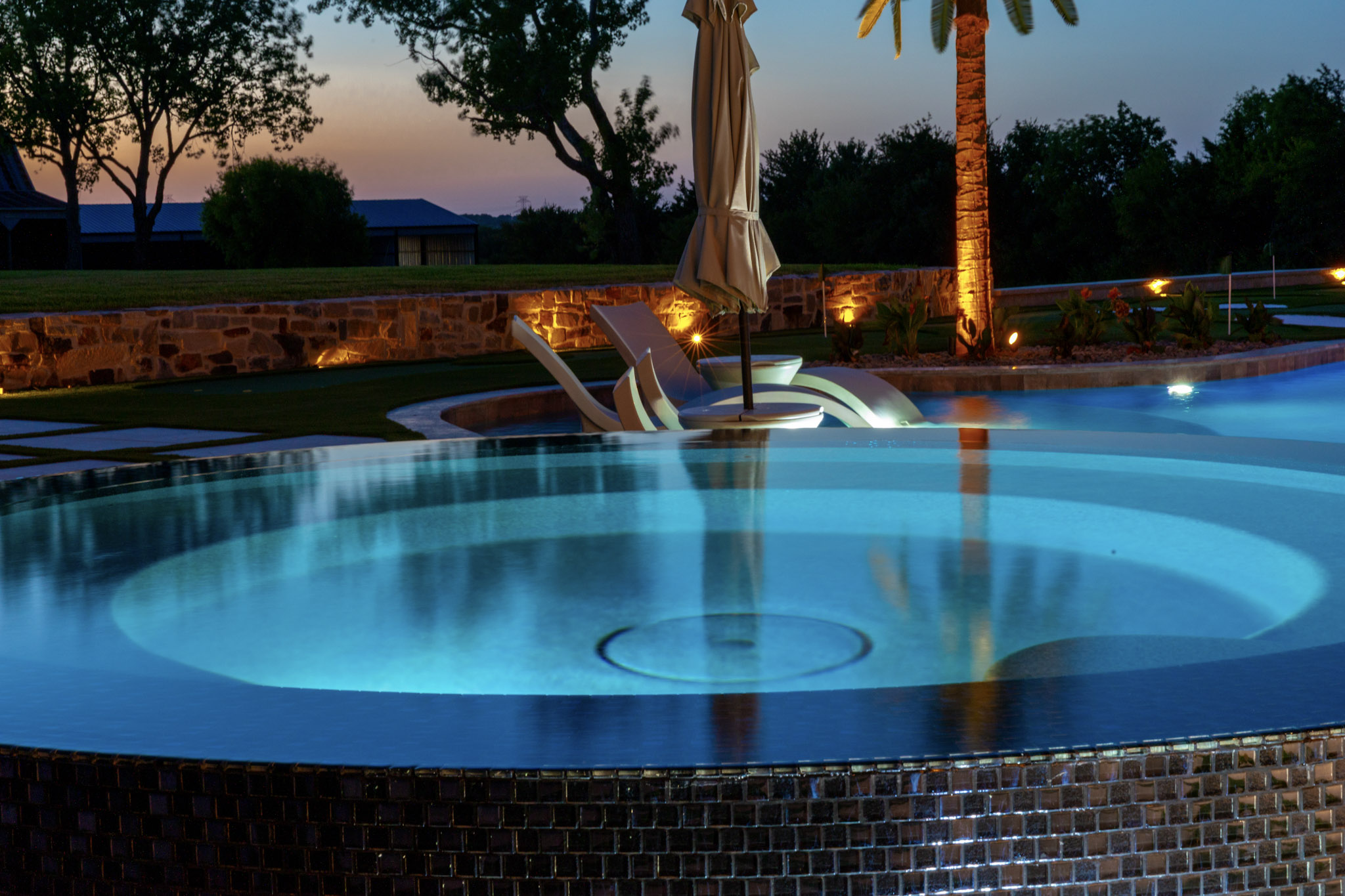 Circular glass-tile spillover spa illuminated blue at twilight.