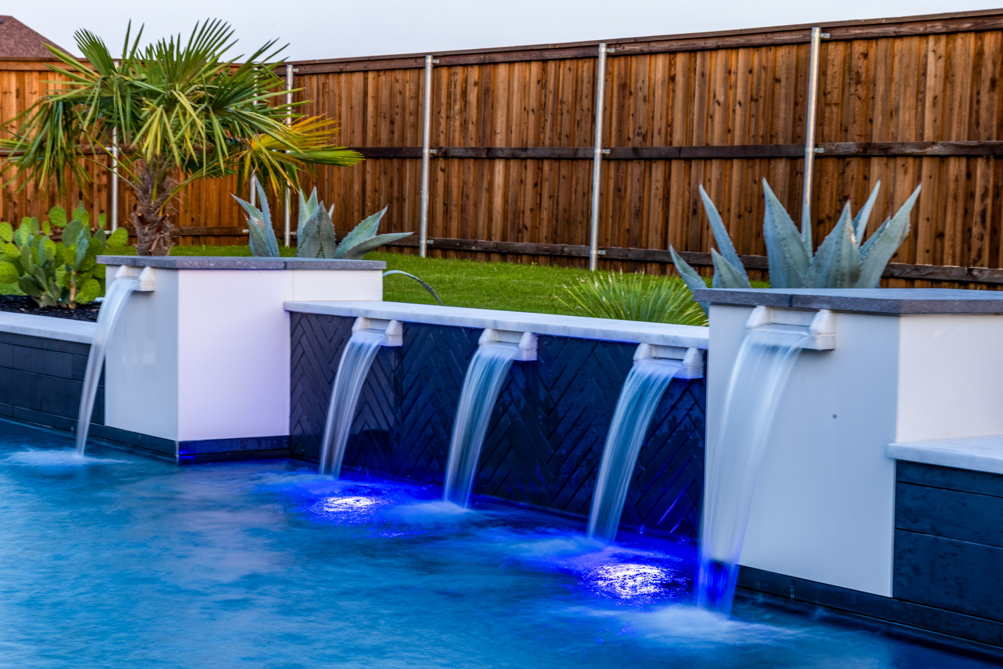 Raised pool wall with five cascading water features, dark tile and tropical landscaping.