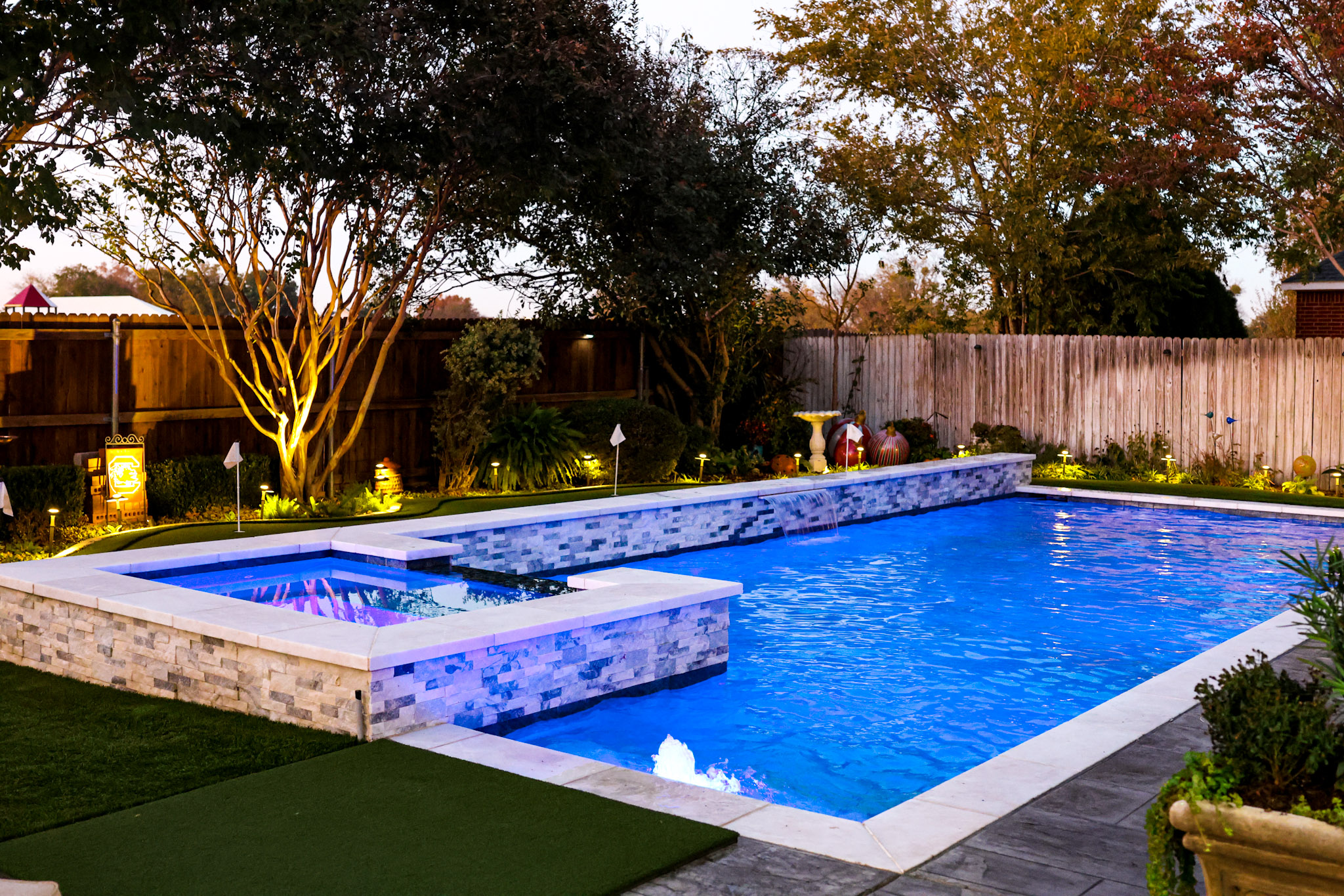Illuminated rectangular pool with raised spa, sheer descent and turf putting green at twilight.