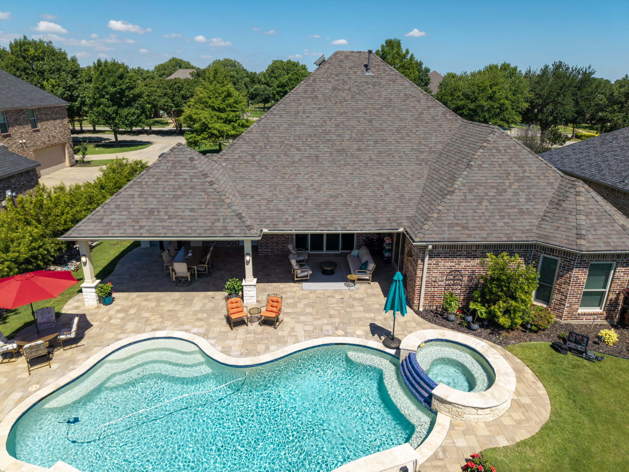 Aerial view of a covered patio behind a brick home with freeform pool, raised spa and paver deck.